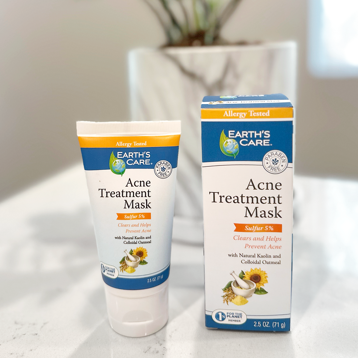 Earth's Care Acne Treatment Mask tube and matching retail box on a light marble countertop, with a blurred potted plant in the background.