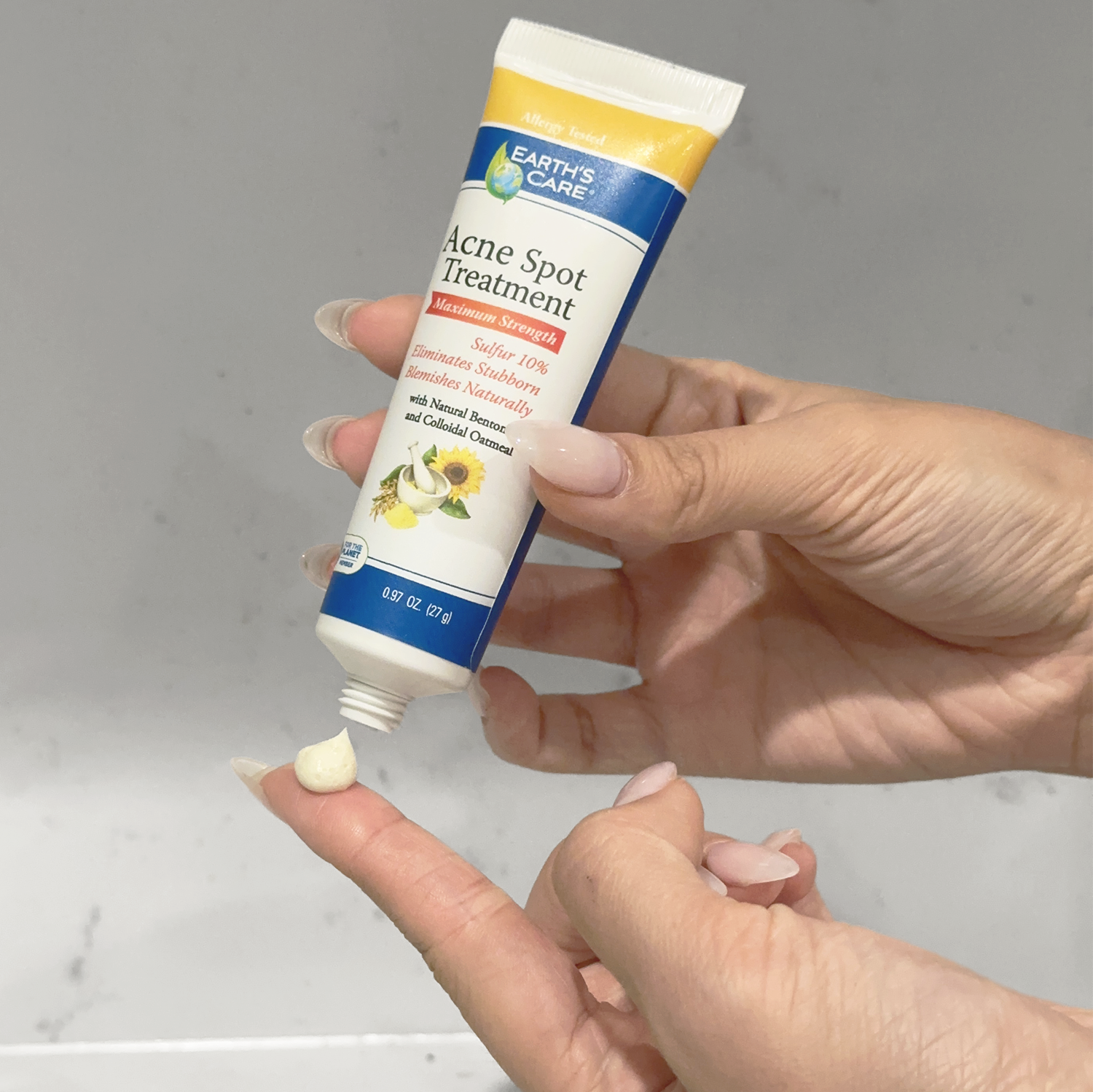 A hand holds an Earth's Care Acne Spot Treatment tube while squeezing a small dollop of white cream onto another finger, against a light gray marble background.