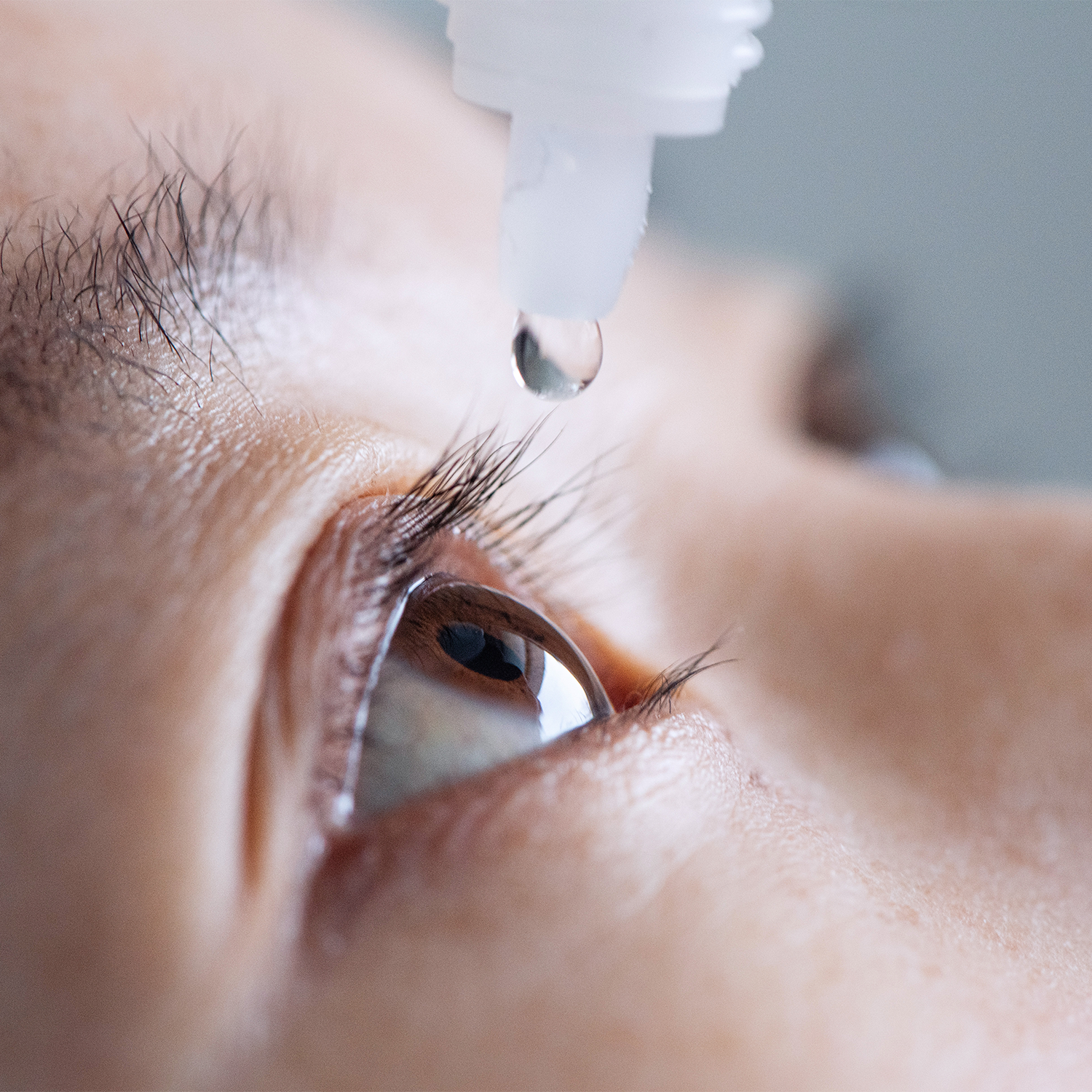 A close-up of a person's eye with a single clear liquid drop hanging from a dropper above the eyelid, showing detailed skin, eyebrow, and iris textures.