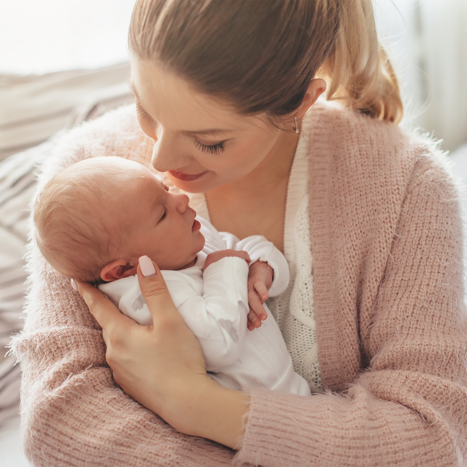A close-up lifestyle shot of a woman in a dusty pink knit cardigan cradling a sleeping newborn baby dressed in a white onesie.