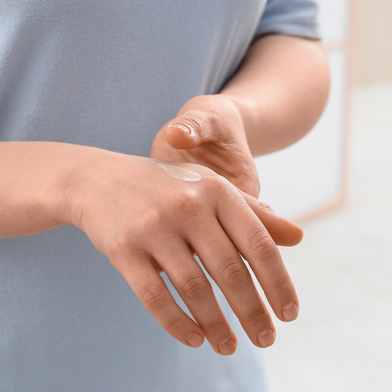 A close-up of a person's hands applying or spreading a clear cosmetic cream or salve on the back of one hand, with a light blue shirt visible.
