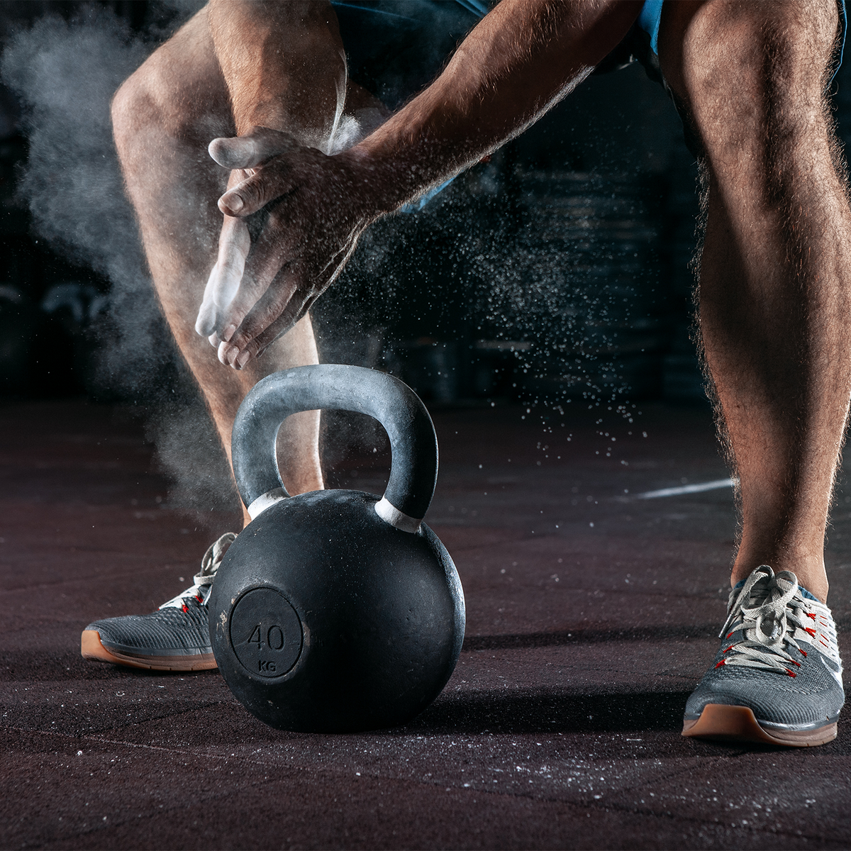 A close-up action shot of a person in athletic wear applying white chalk to their hands while holding a black kettlebell on a dark gym floor. The person's legs and hands are visible, with chalk dusting in the air.