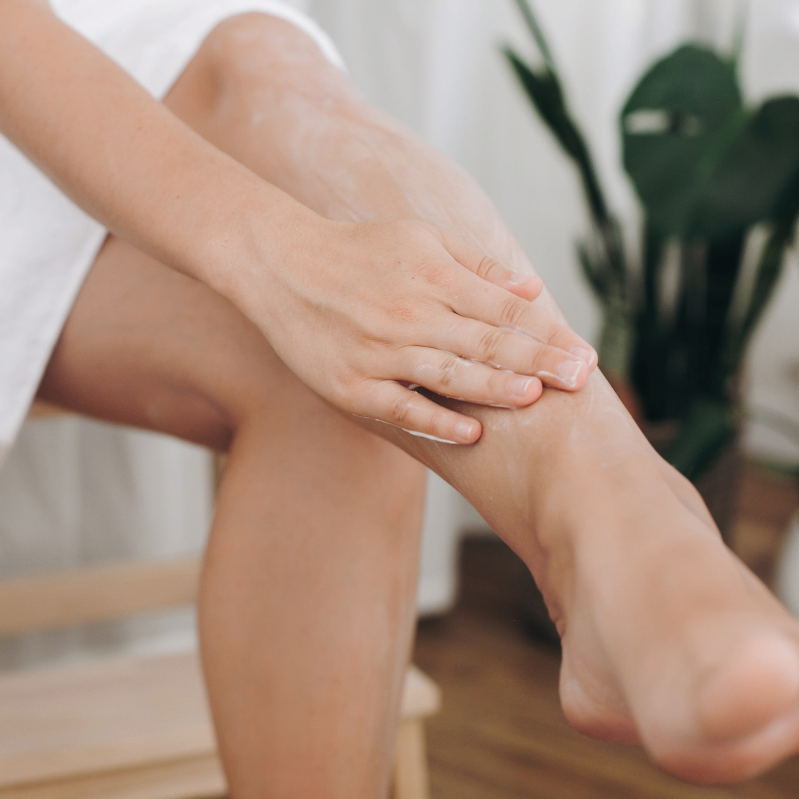 A close-up lifestyle shot of a person applying a white lotion or cream onto their lower leg and ankle, with a blurred indoor background featuring a green plant.