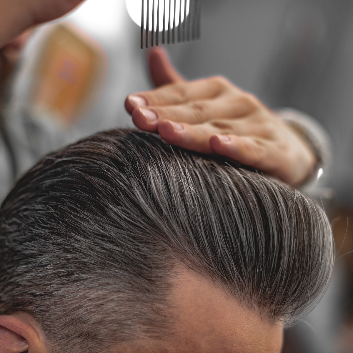 Close-up of a person with styled hair, showing the top of the head and side hair with a hand combing through the hair. A hand is visible at the top holding a comb.