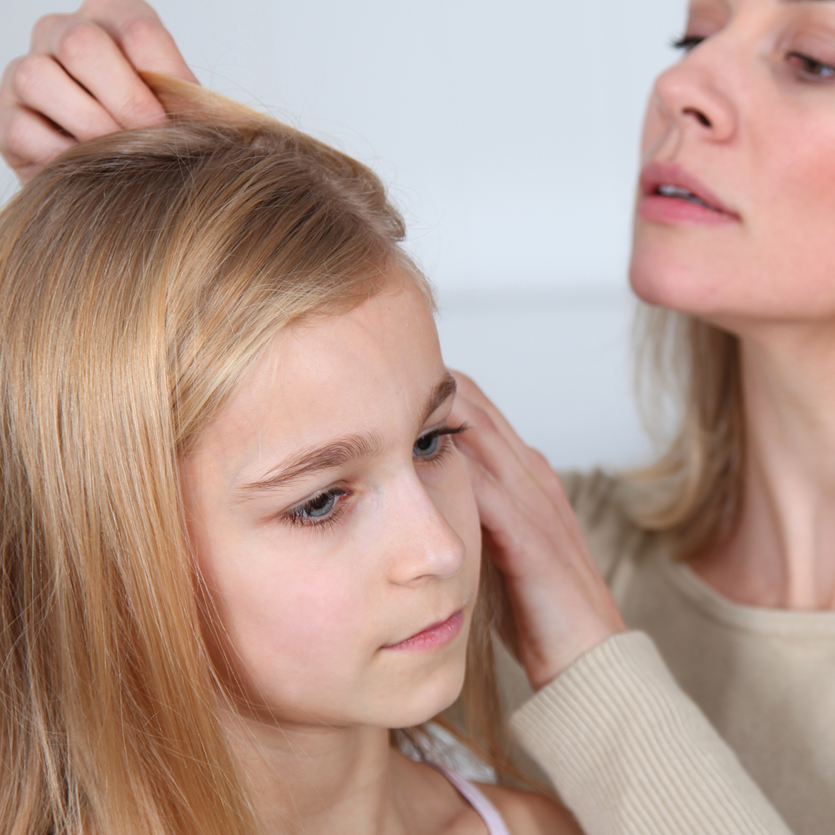 A woman inspecting the hair of a young blonde girl, with the girl wearing a light pink tank top against a soft light gray background.