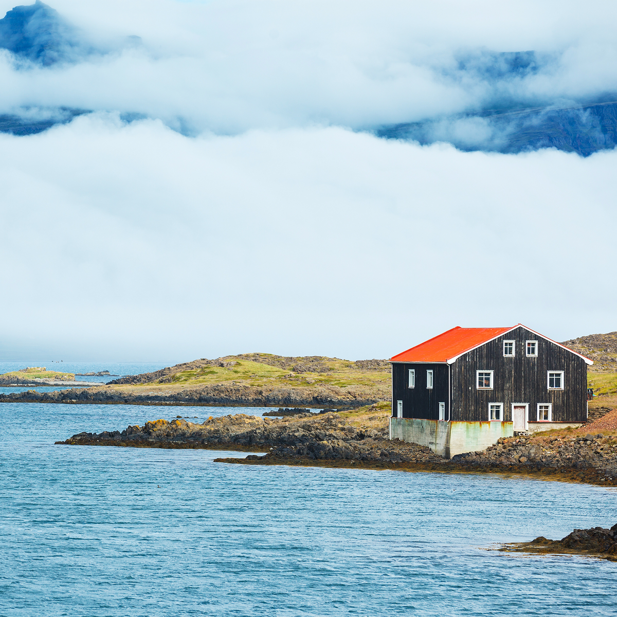 A small black wooden house with a bright orange-red roof sits on a rocky shoreline beside calm blue water, with grassy hills and cloudy mountains in the background.