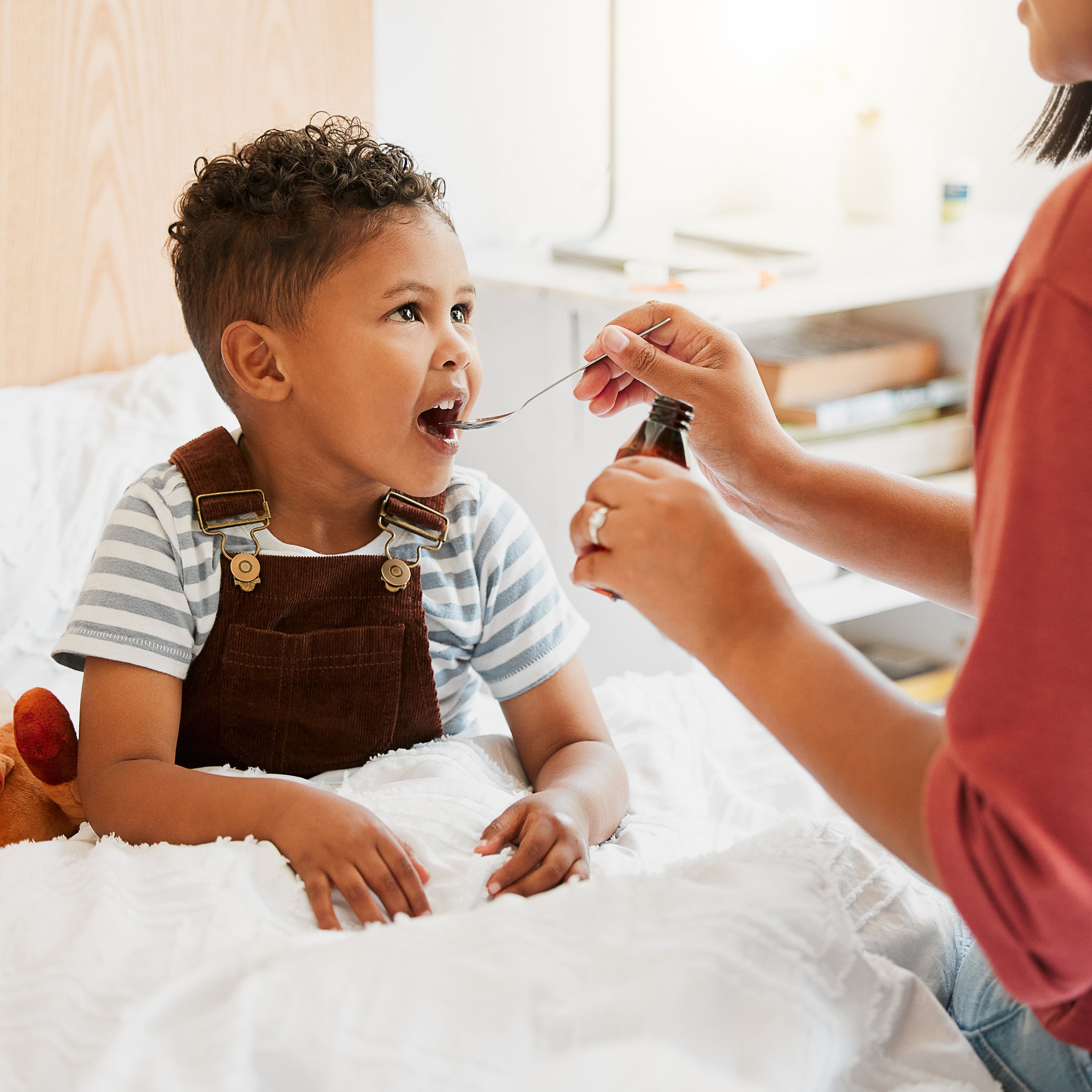 A young child wearing brown overalls and a striped shirt sits on a bed while an adult in a coral-red top administers medicine from a small bottle using a spoon. A plush toy rests beside the child on white bedding in a bright bedroom.