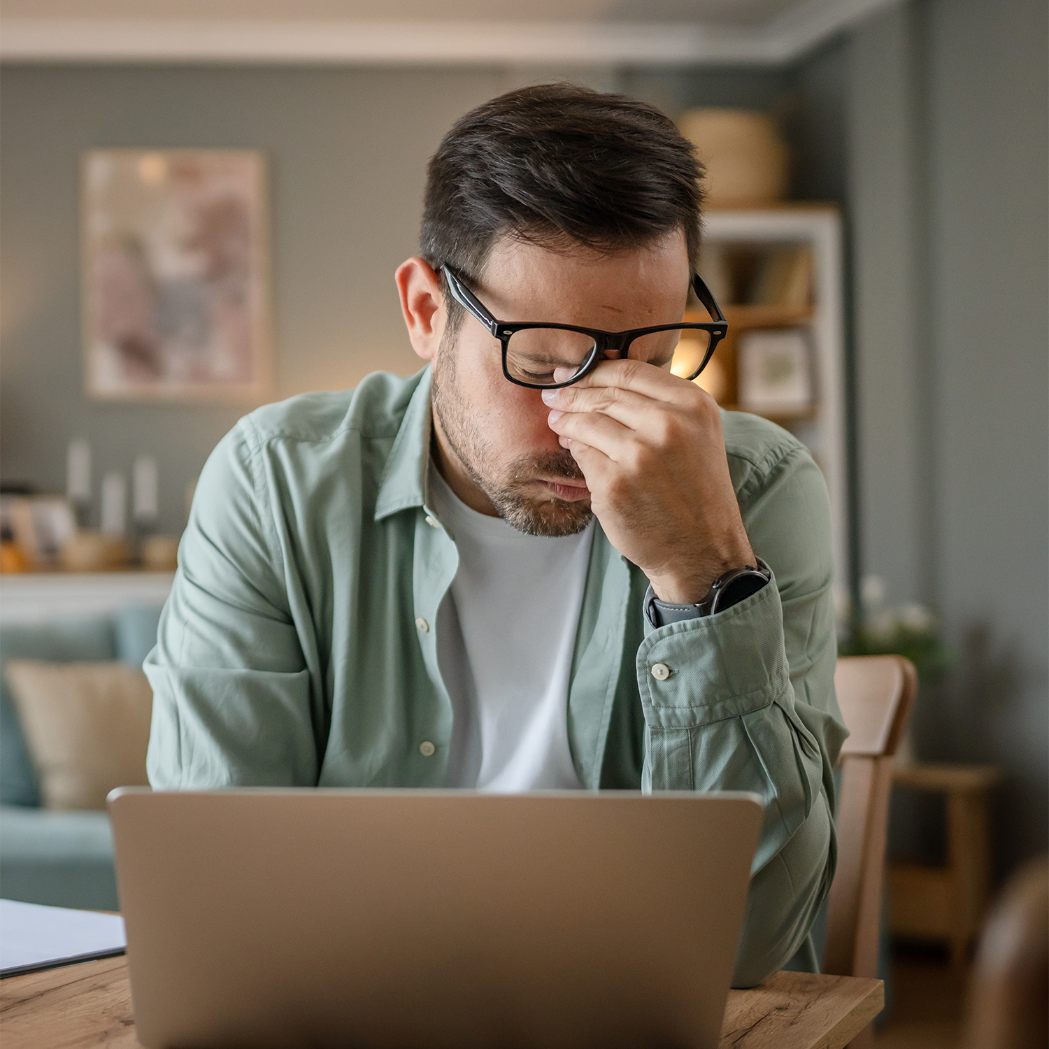 A man wearing black-rimmed glasses and a sage green button-up shirt sits at a wooden table in a home office, rubbing his eyes while looking at a laptop.