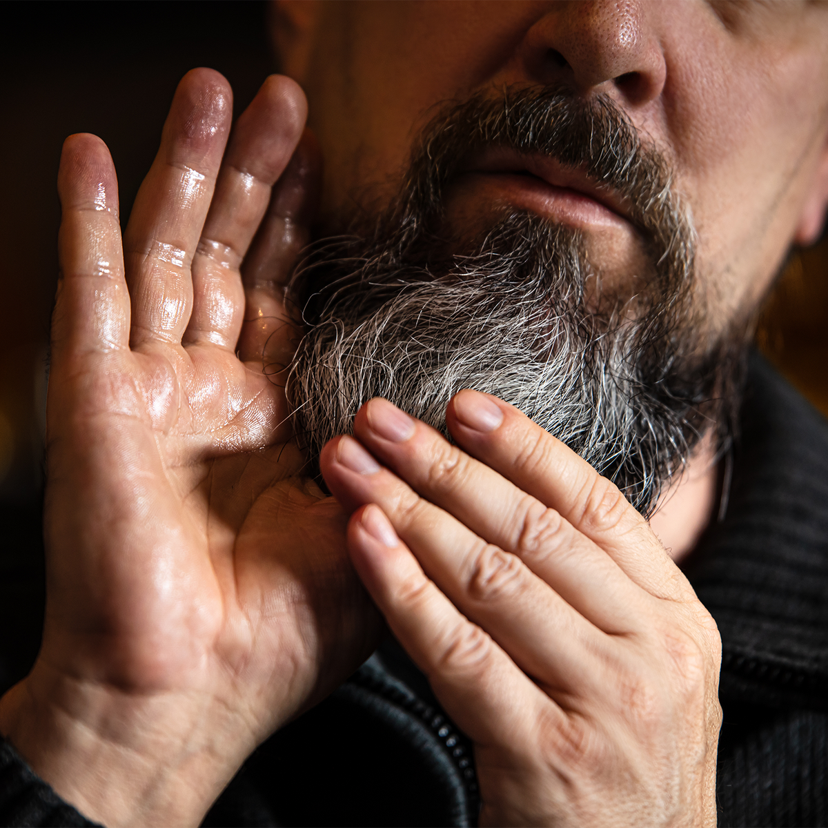 A close-up of a man's hands and beard, showing a glossy, shiny hair or beard oil being applied. The product is glistening on his fingers and chin, highlighting its moisturizing and conditioning properties.