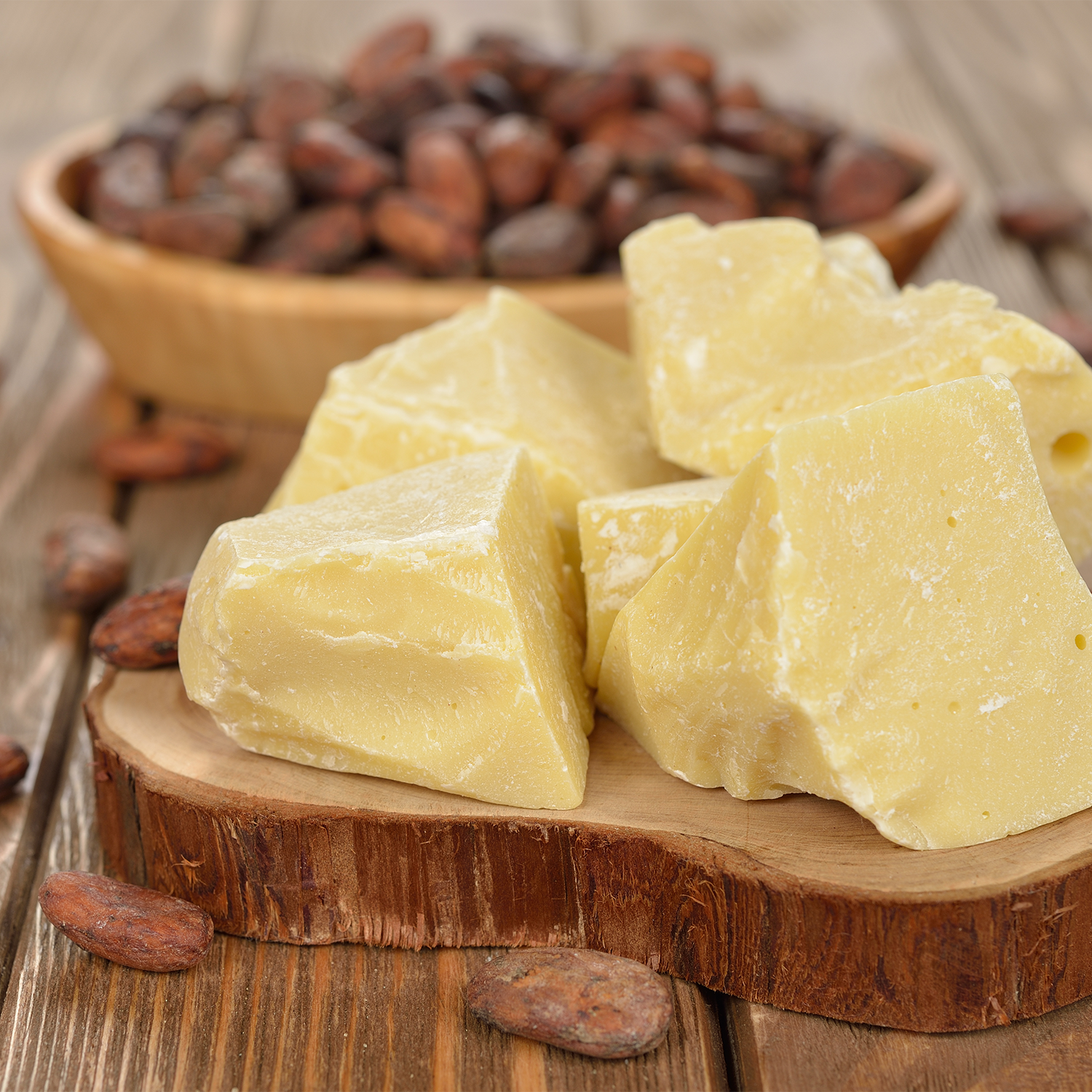 Blocks of pale yellow shea butter arranged on a wooden slice stand, with a bowl of cocoa beans and scattered cocoa beans on a rustic wooden table.