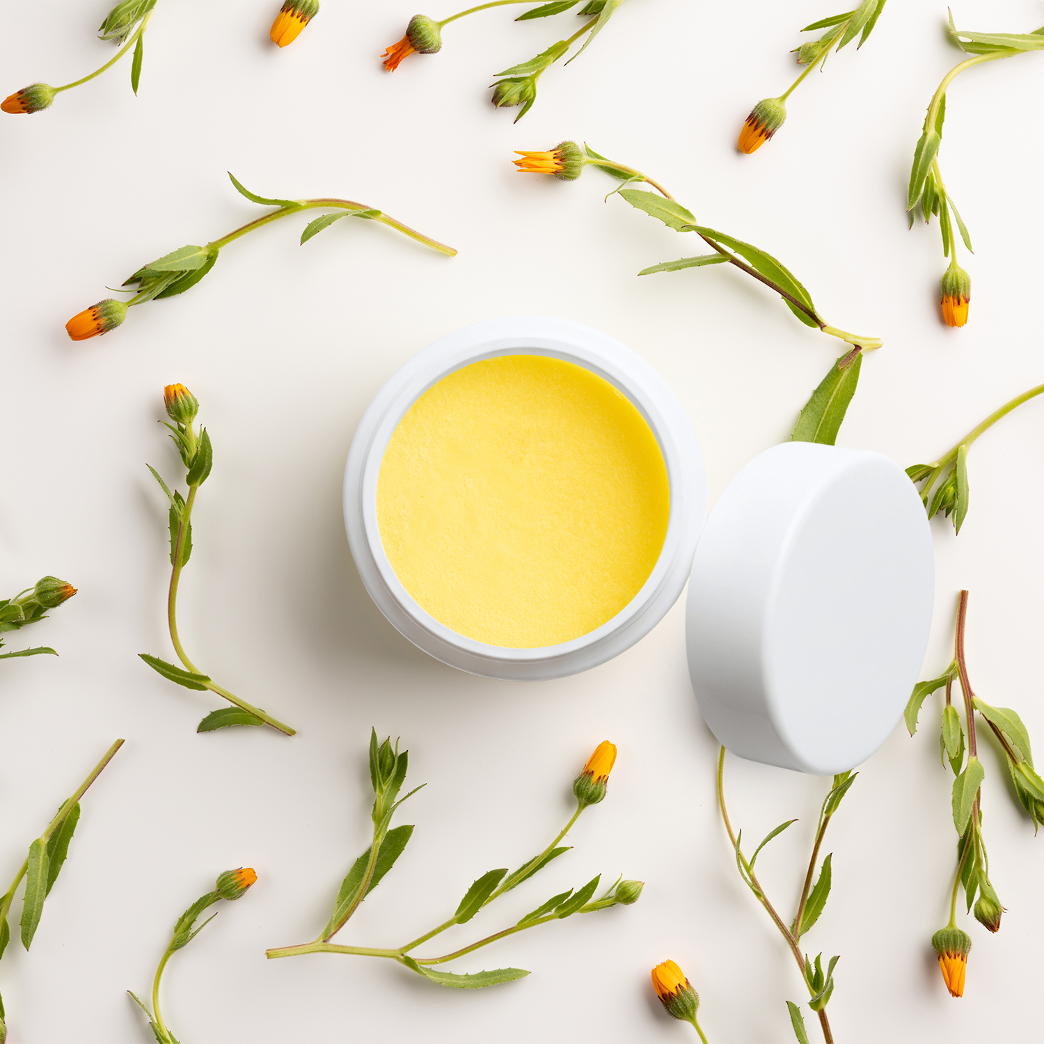 A white cosmetic jar filled with bright yellow balm or cream, shown open with the lid beside it on a light background, surrounded by scattered green plant stems and small orange flower buds.