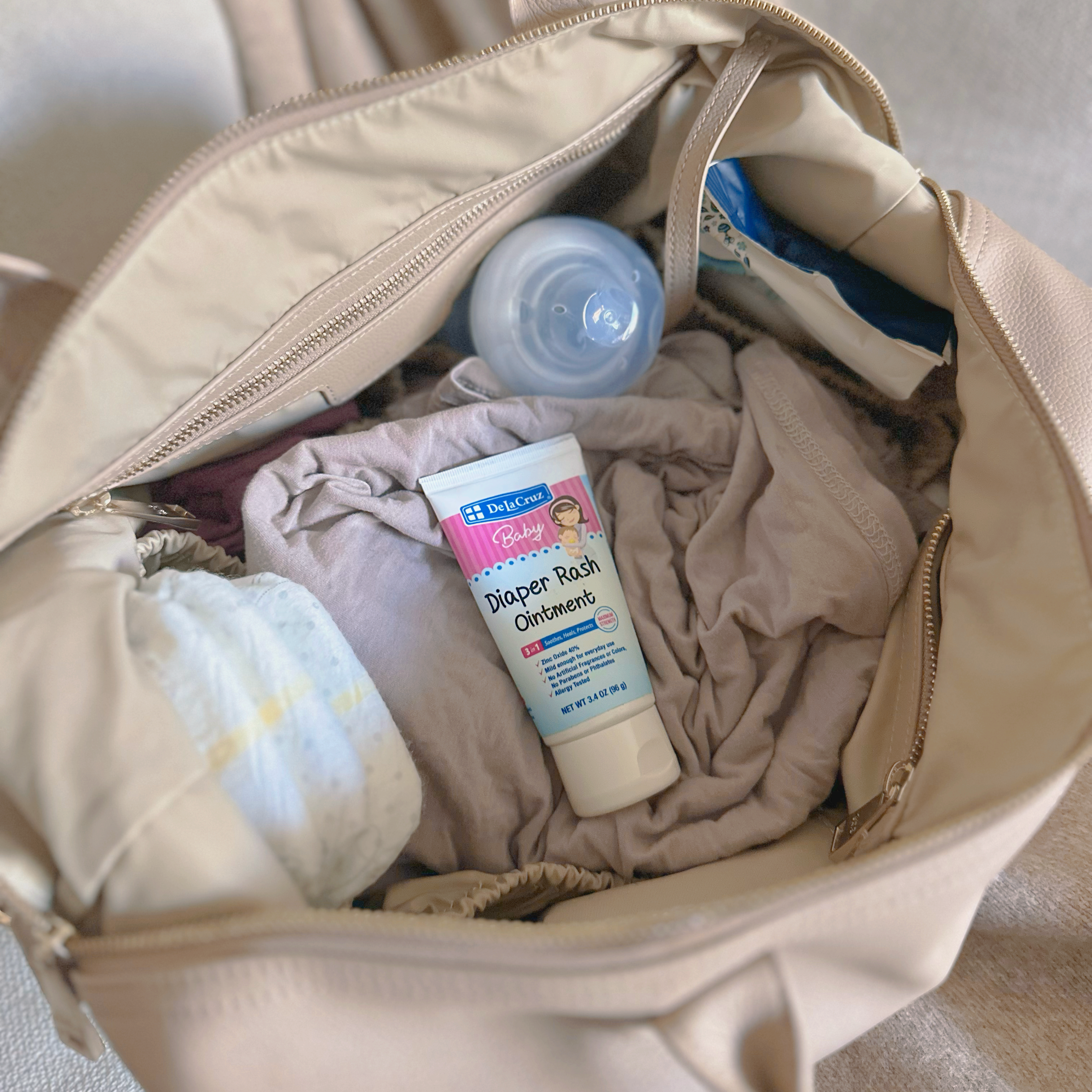An open beige diaper bag filled with baby essentials, including a diaper, a tube of diaper rash cream, a bottle cap, and folded cloth items, photographed on a soft neutral fabric background.