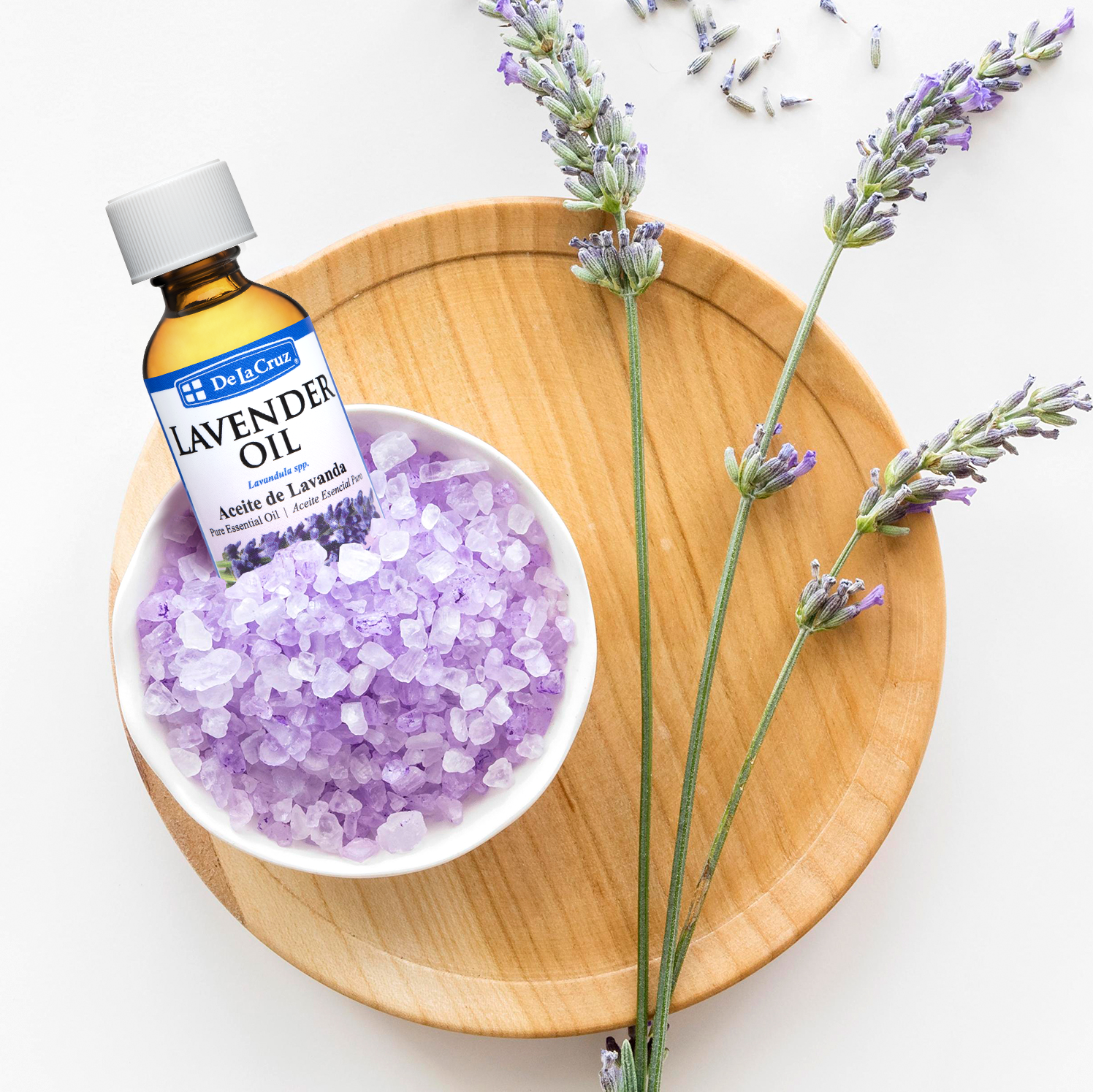 A bottle of De La Cruz Lavender Oil placed beside a white bowl filled with lavender crystals on a round wooden tray, with lavender sprigs arranged on a white background.