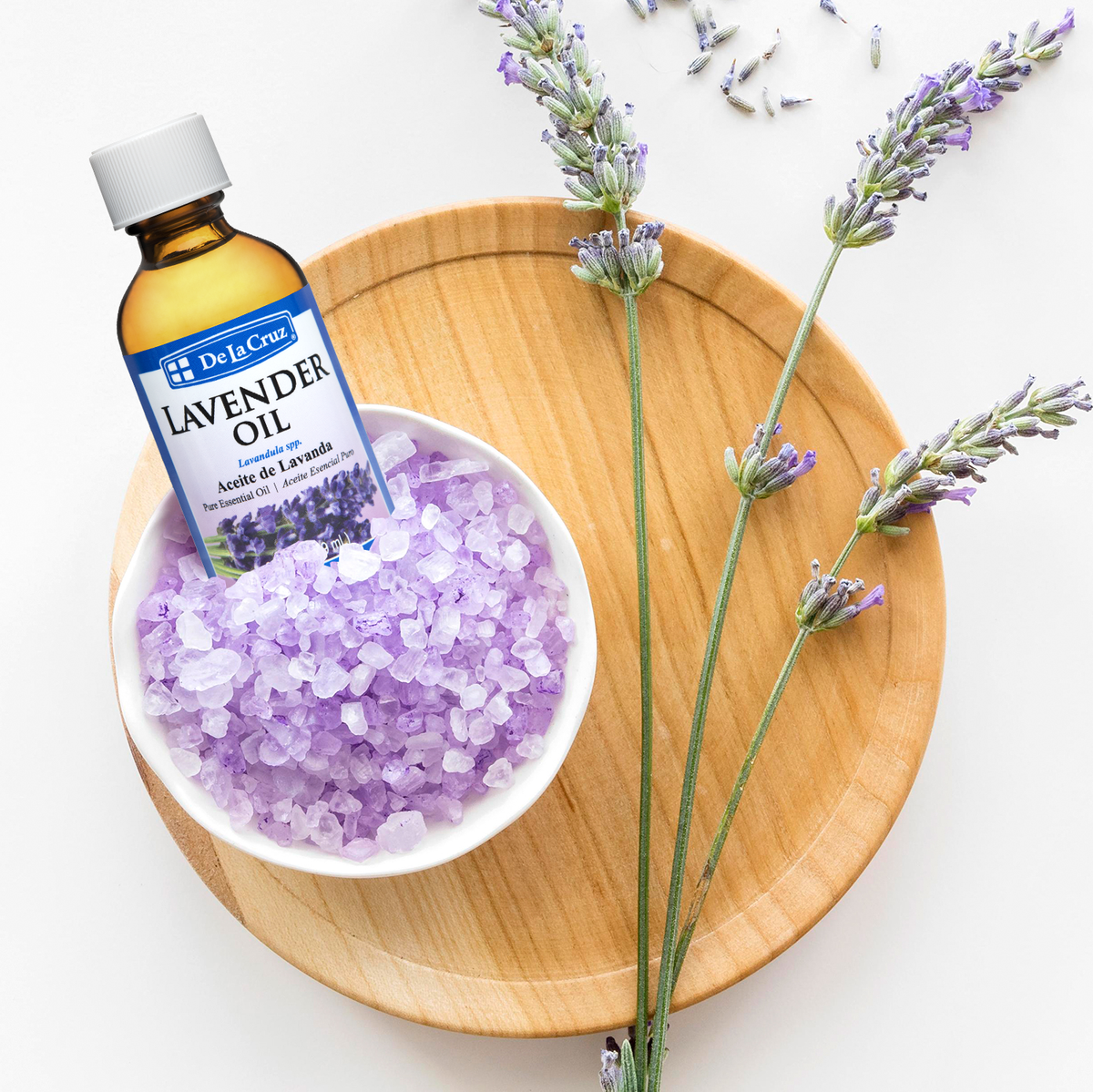 A bottle of De La Cruz Lavender Oil placed in a white bowl filled with lavender bath salts, styled on a round wooden tray with lavender sprigs on a white background.
