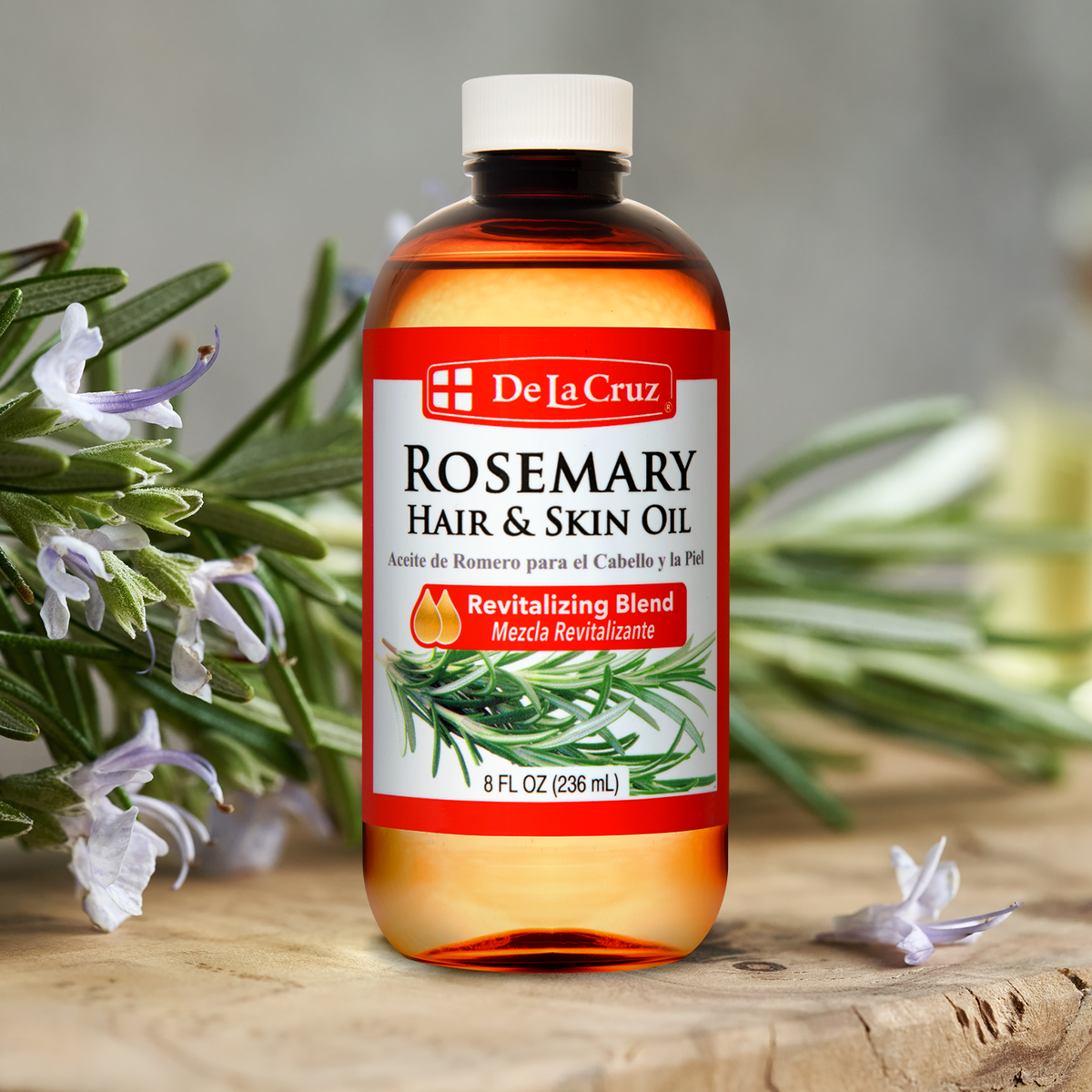 A bottle of De La Cruz Rosemary Hair & Skin Oil stands on a rustic wooden surface with rosemary sprigs and small white flowers around it. The bottle is amber-colored with a white cap and a red-and-white label featuring rosemary imagery.