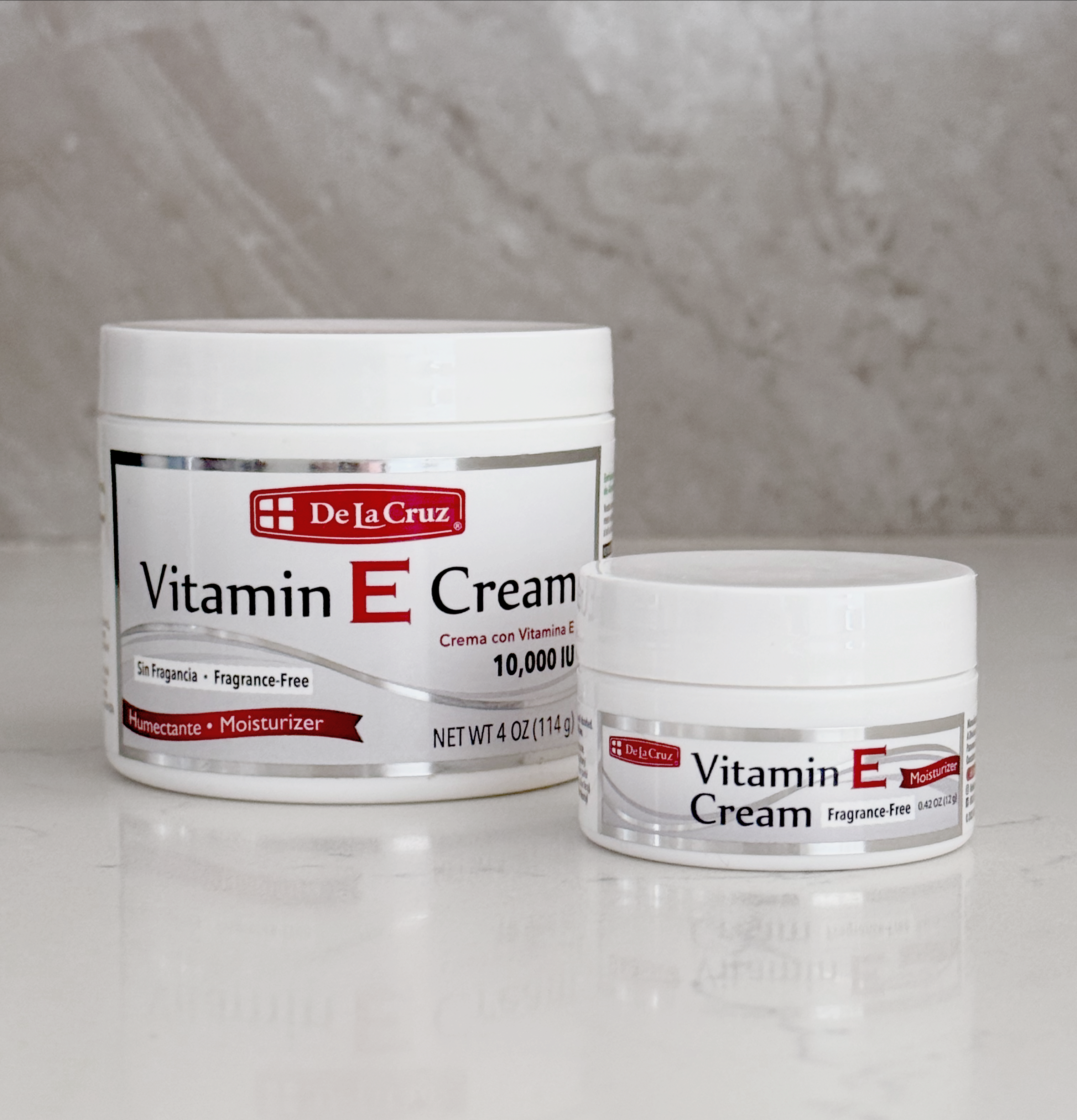 Two white jars of De La Cruz Vitamin E Cream on a reflective white surface against a gray marble-textured background.