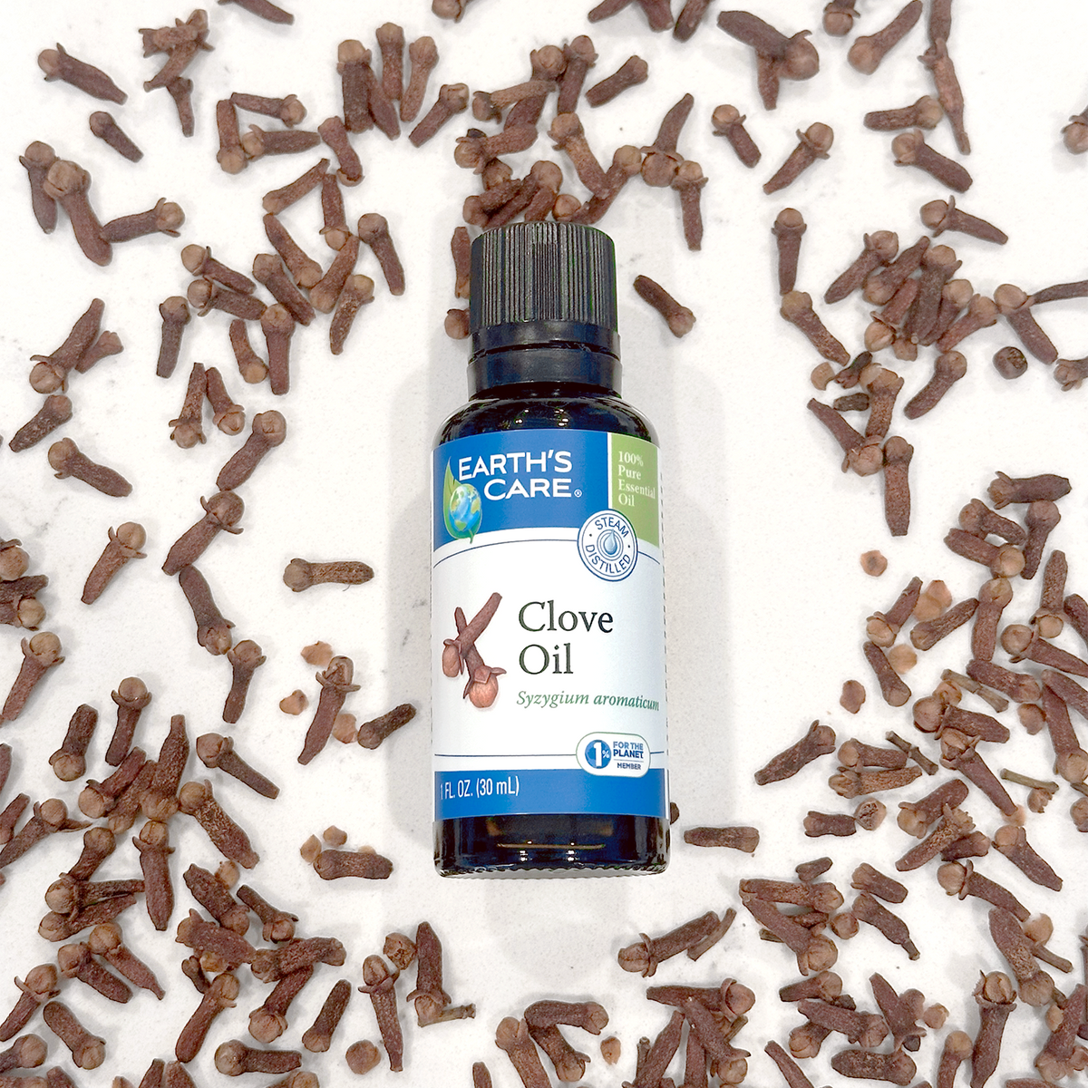 A bottle of Earth's Care Clove Oil centered on a white marble surface, surrounded by scattered brown cloves. The bottle has a blue and white label with a black cap.