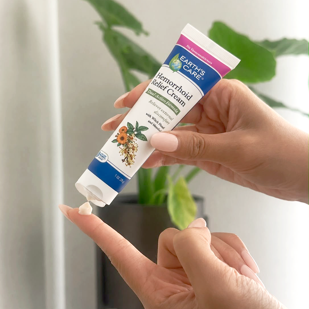 A person's hands holding and squeezing a tube of Earth's Care Hemorrhoid Relief Cream, with a small dollop of the white cream being dispensed onto another finger, set against a soft-focus background with green plants.