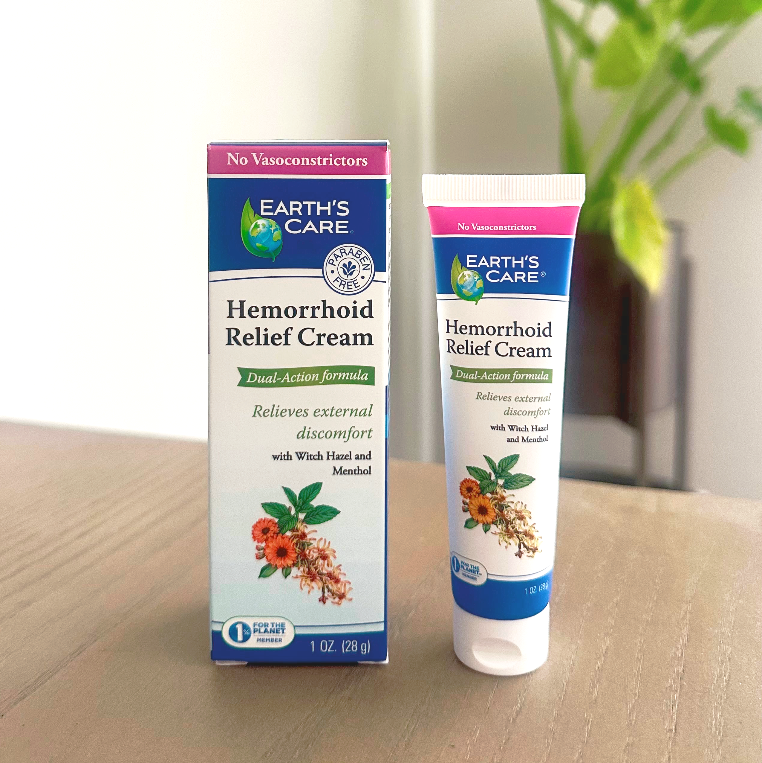 Earth's Care Hemorrhoid Relief Cream shown as a white squeeze tube with blue and pink branding next to its matching cardboard box, placed on a light wooden tabletop with a blurred indoor background.