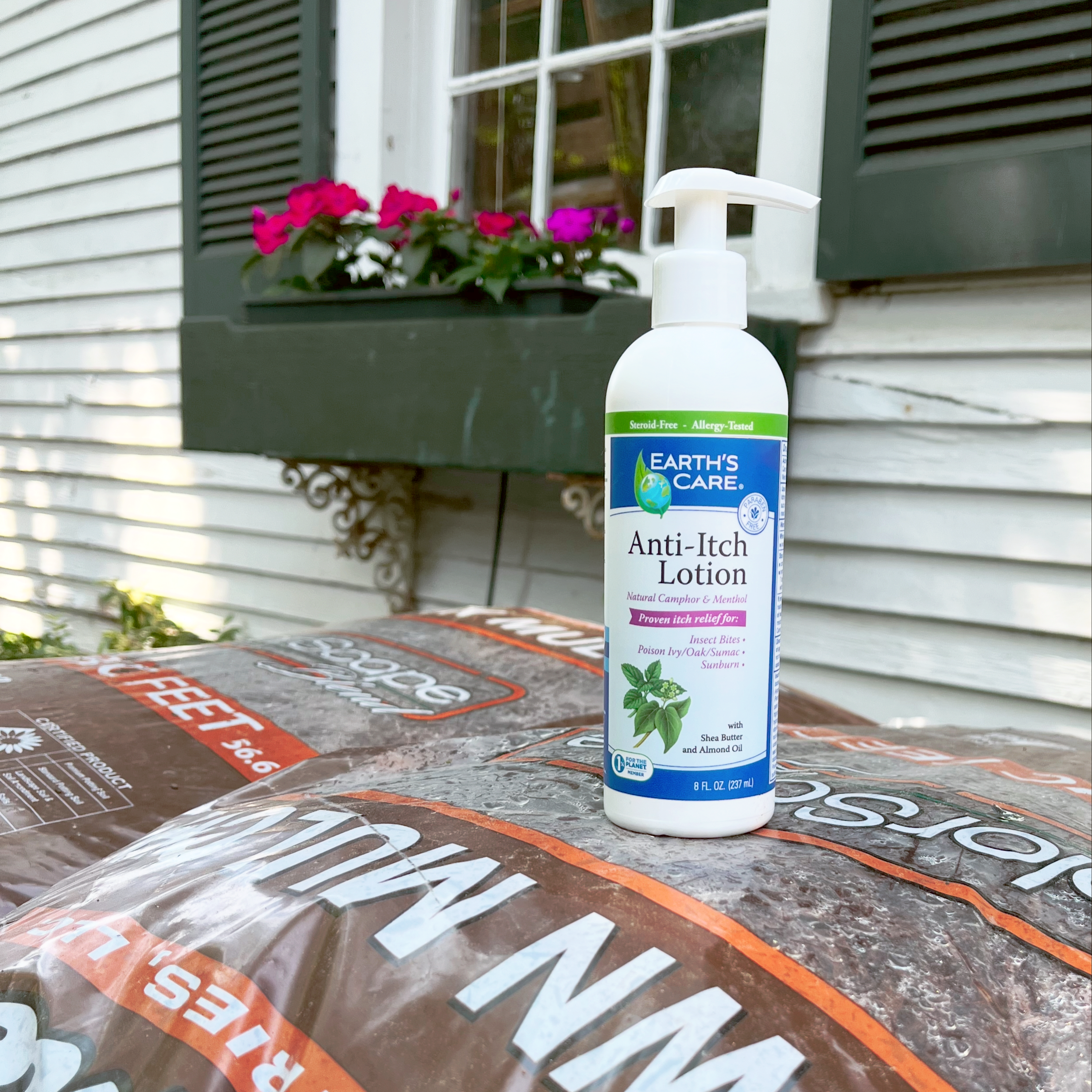 Earth's Care Anti-Itch Lotion bottle placed on top of a large bag of lawn treatment in front of a house exterior with white siding, dark green shutters, and pink flowers in a window box.