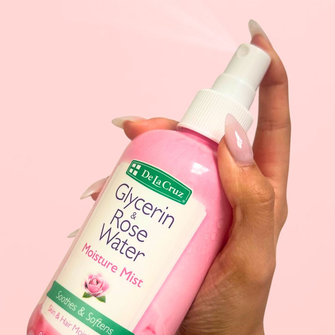 A hand holding a pink spray bottle of De La Cruz Glycerin & Rose Water Moisture Mist against a solid pink background.
