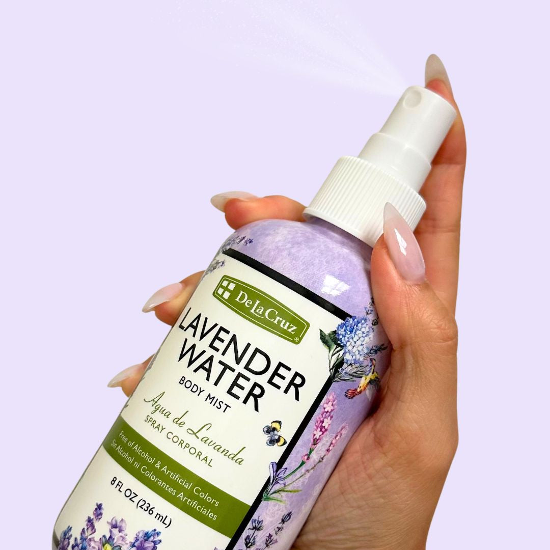 A hand holding a bottle of De La Cruz Lavender Water Body Mist against a solid light lavender background, with the spray nozzle emitting a fine mist.