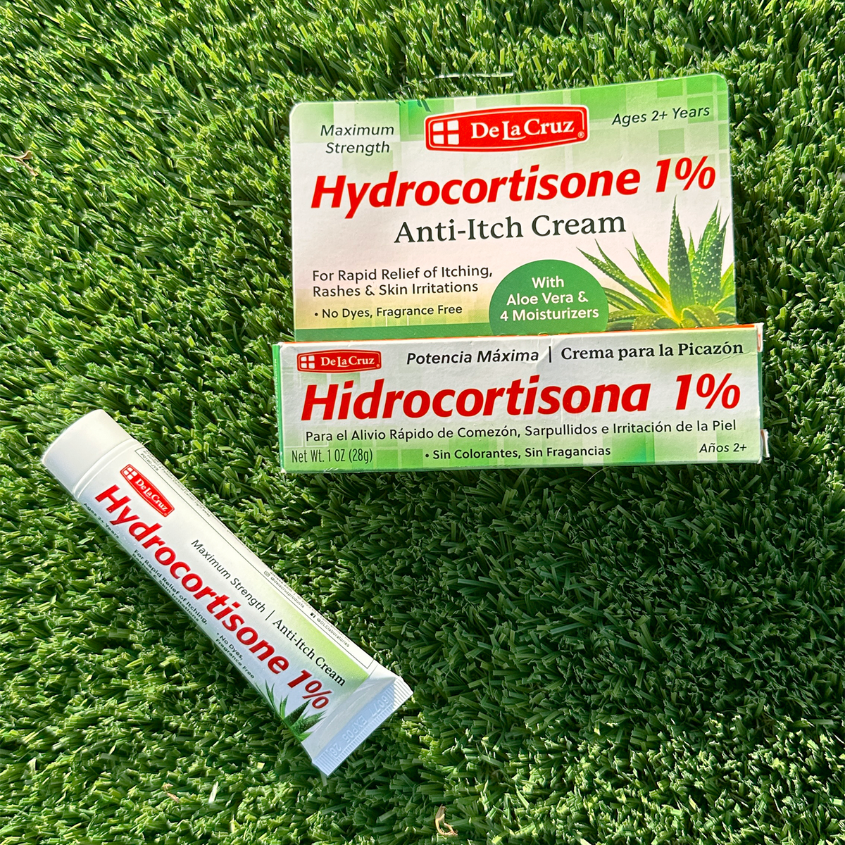 A tube and two boxes of De La Cruz Hydrocortisone 1% Anti-Itch Cream arranged on a green grass background.