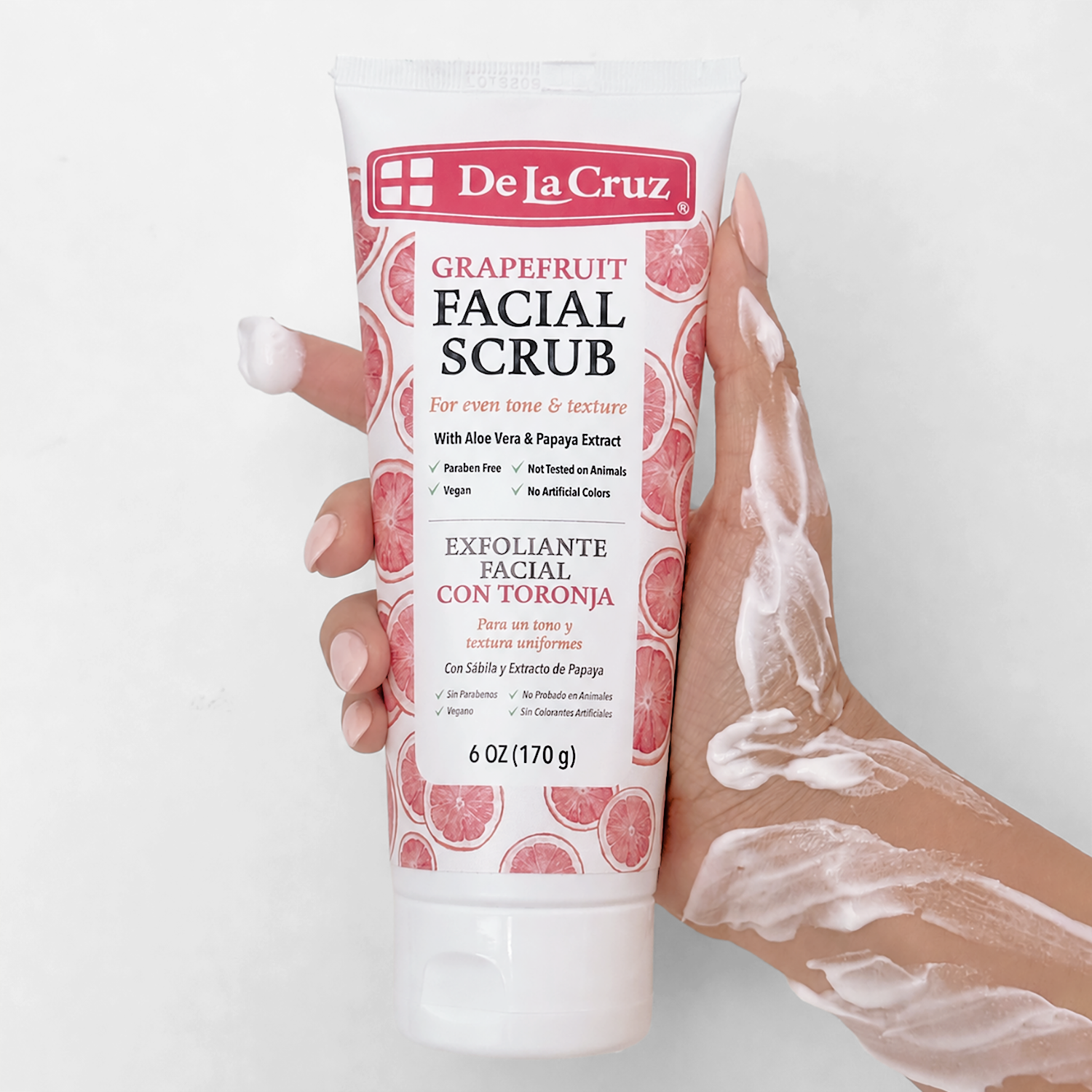 A hand holds a tube of De La Cruz Grapefruit Facial Scrub, with white cream applied to the fingers and wrist, against a clean white background.