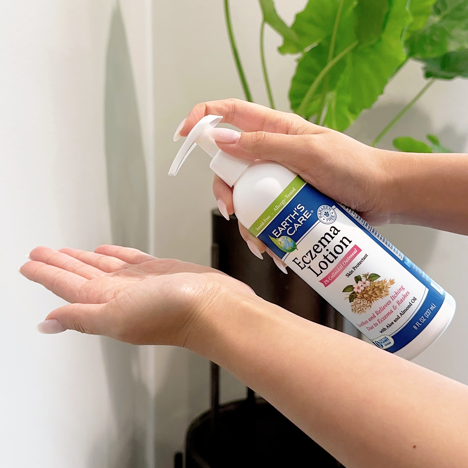 A person's hands holding and dispensing Earth's Care Eczema Lotion on another hand, with a green plant in the background.