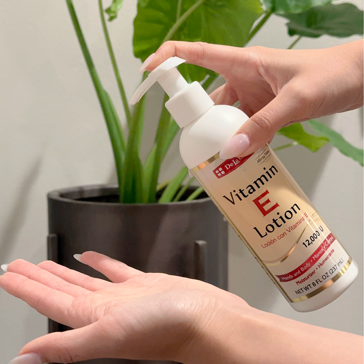 Hand holding De La Cruz Vitamin E Lotion with green plant in the background. 