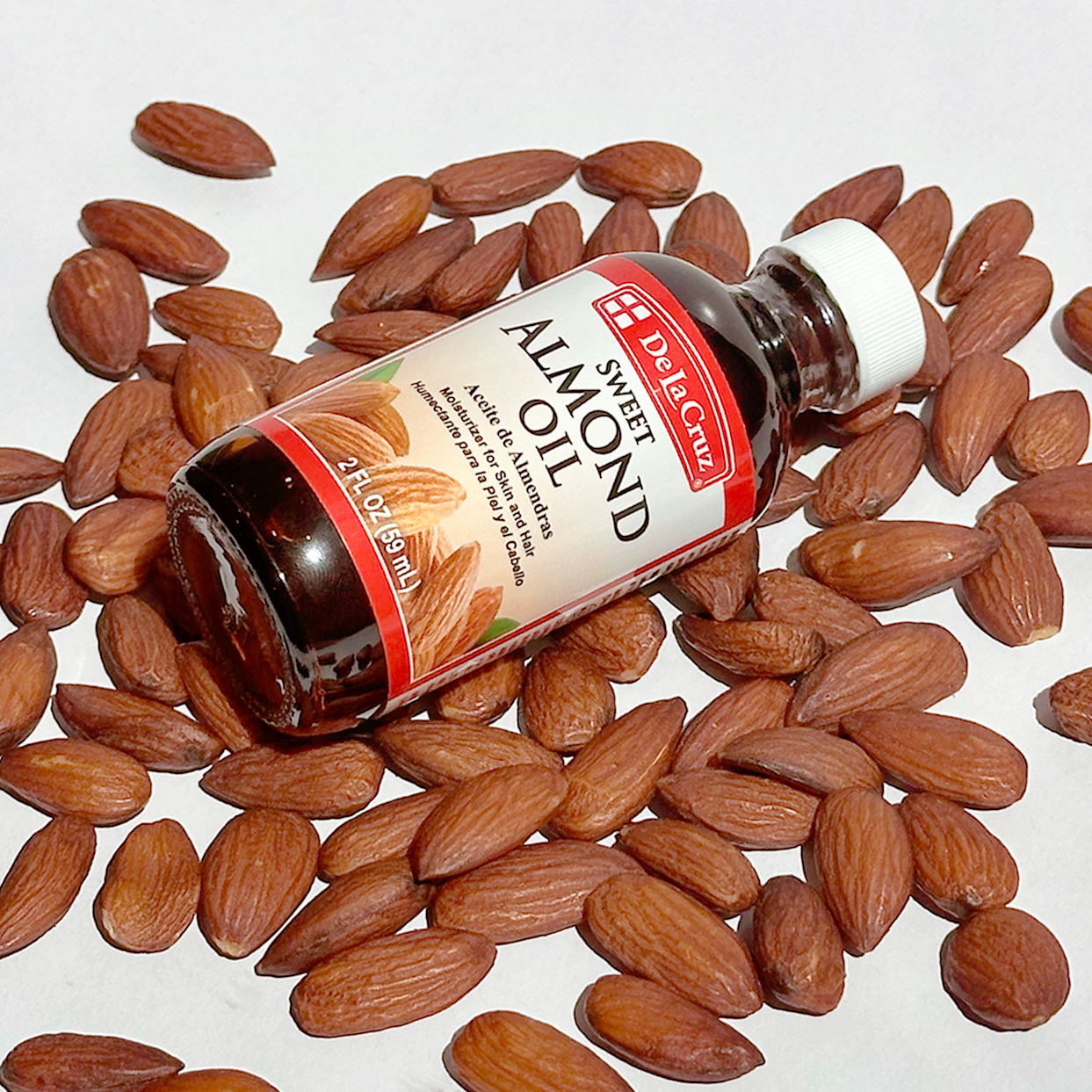 A bottle of De La Cruz Sweet Almond Oil lies on its side among scattered whole almonds on a plain white background.