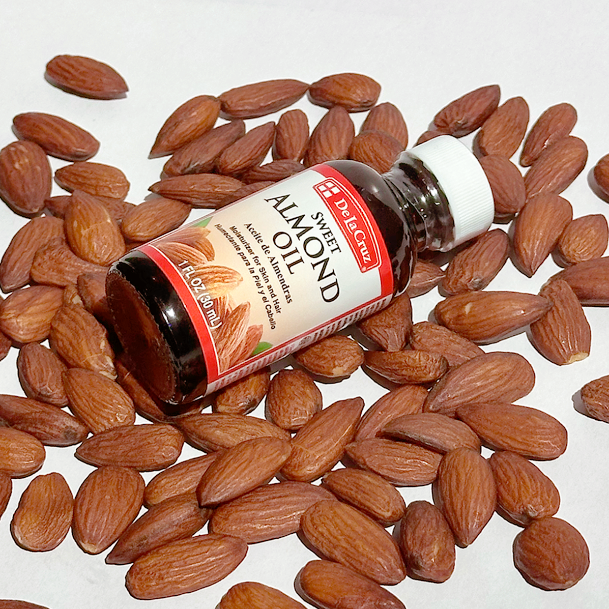 A small bottle of De La Cruz Sweet Almond Oil lies on its side among scattered whole almonds on a plain white background.