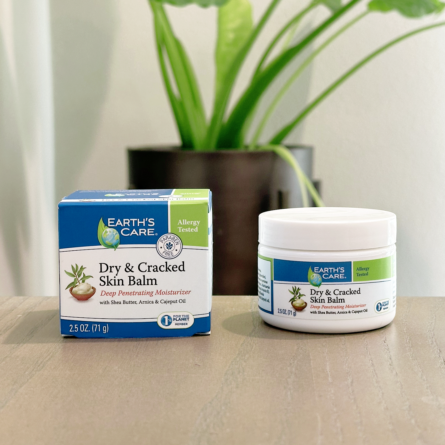 Earth's Care Dry & Cracked Skin Balm shown with its blue and white retail box and a white plastic jar on a light wood surface, with a blurred green potted plant in the background.