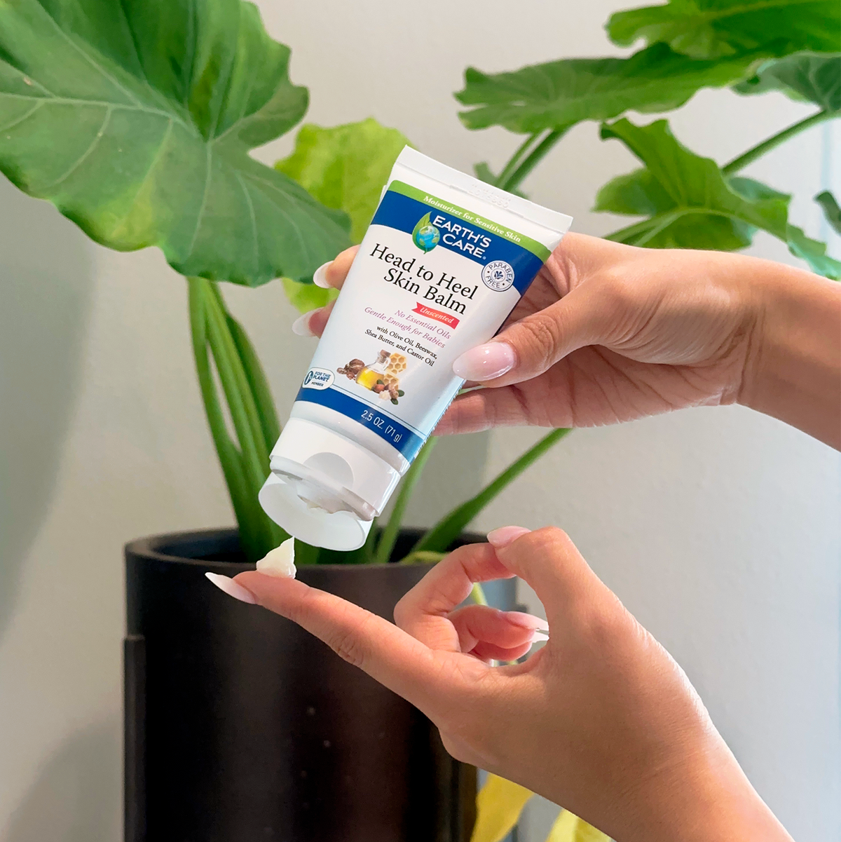A person's hands are shown squeezing Earth's Care Head to Heal Skin Balm onto a fingertip, with green houseplant leaves in the background.