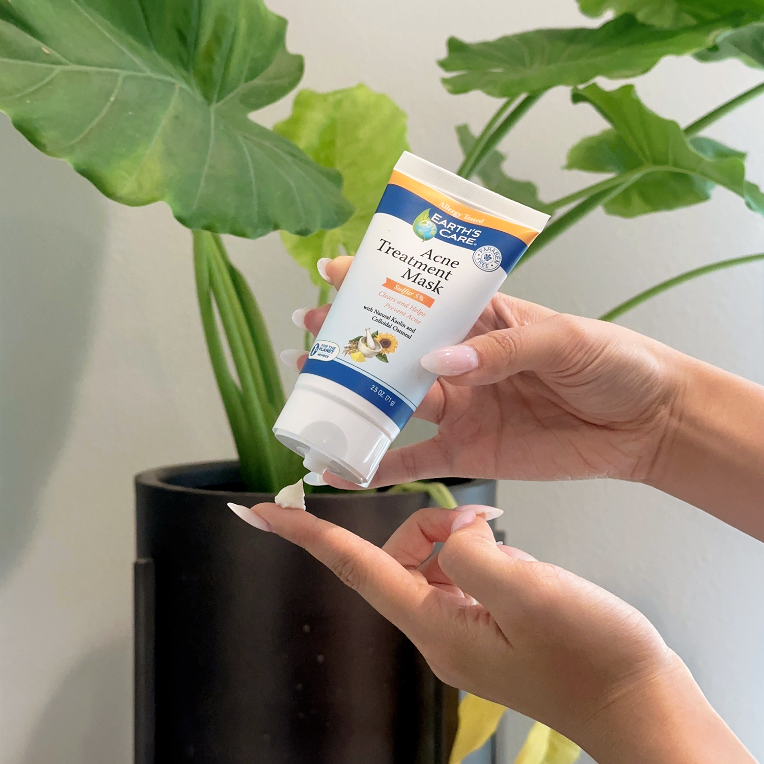 A person's hands are shown squeezing a tube of Earth's Care Acne Treatment Mask onto a fingertip, with a large green potted plant in the background.