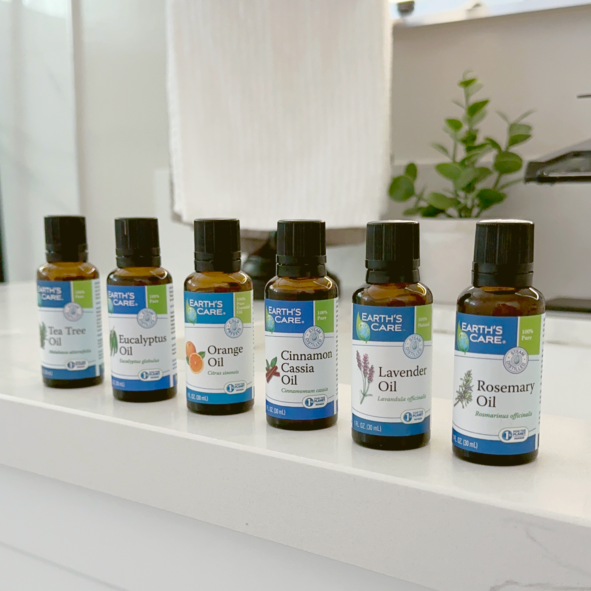 Five small amber glass essential oil bottles with black caps lined up on a white bathroom counter. The labels show different scents including Bergamot, Cinnamon, Lavender, and Rosemary, with a white towel and bathroom background.