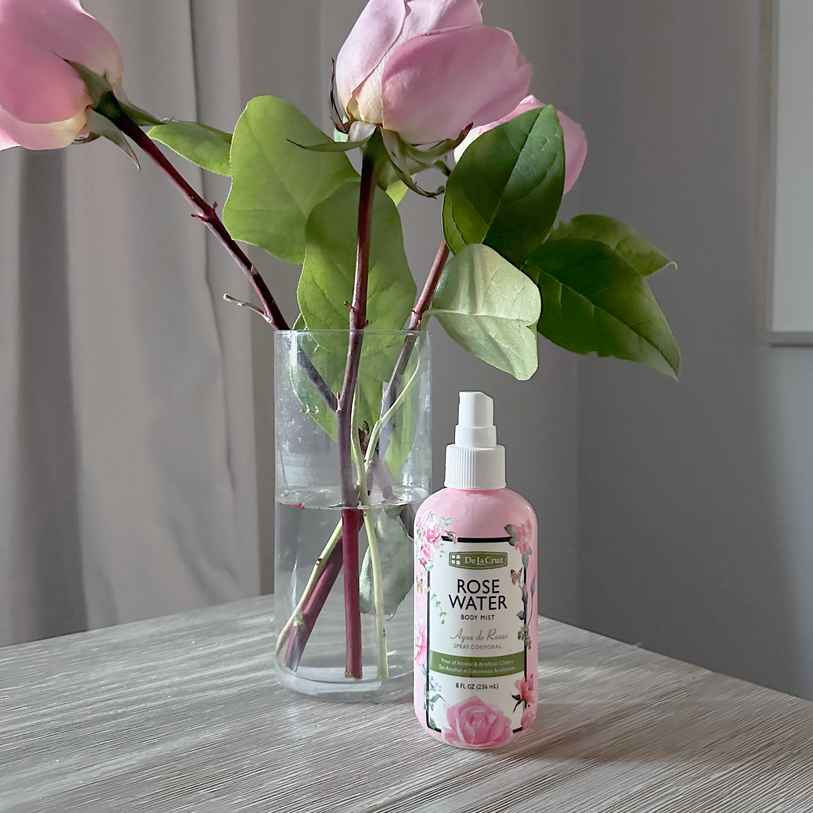 A pink spray bottle labeled Rose Water sits on a light wood table next to a clear glass vase holding pink rose buds and green leaves, in a softly lit indoor setting.