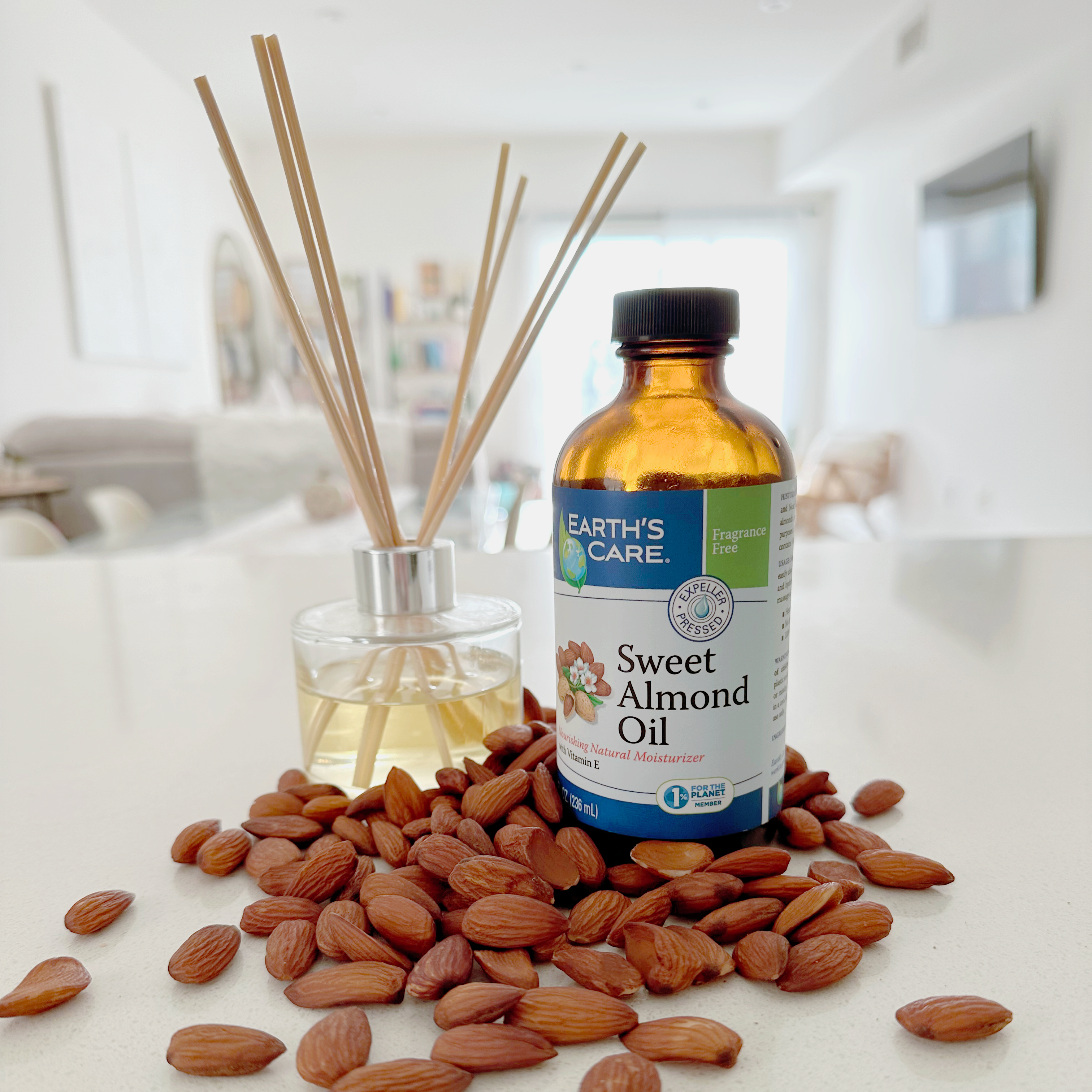 A bottle of Earth's Care Sweet Almond Oil stands on a light countertop surrounded by scattered almonds, with a reed diffuser bottle behind it in a softly blurred indoor setting.