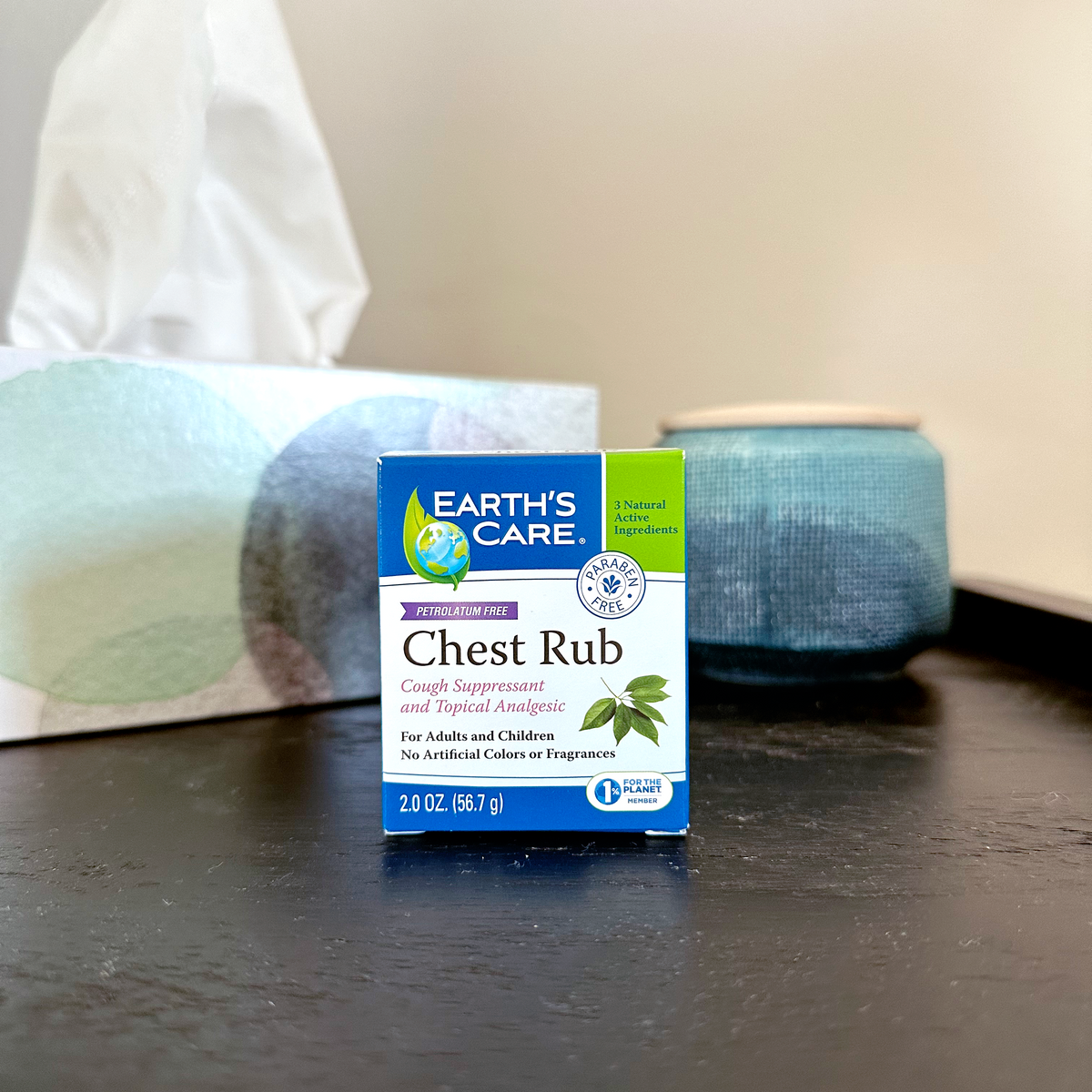 A box of Earth's Care Chest Rub antiseptic and deodorant spray placed on a dark tabletop, with a tissue box and a blue container blurred in the background.