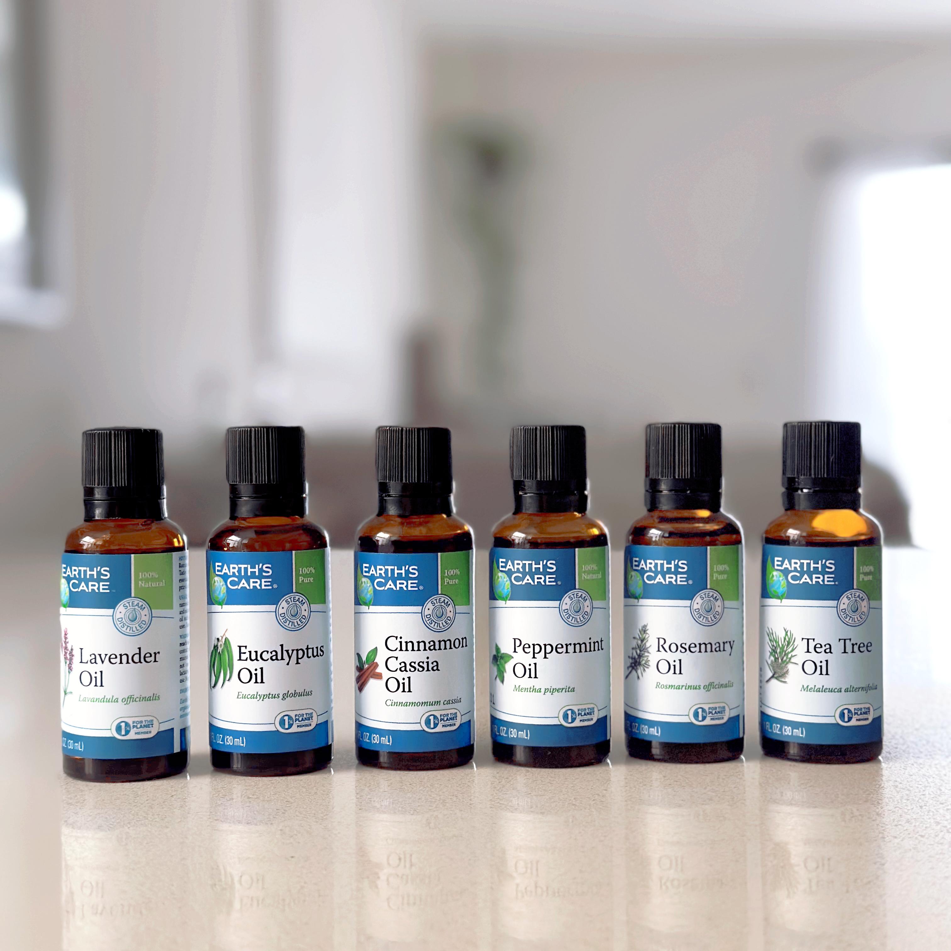 Six amber glass essential oil bottles from Earth's Care lined up on a light countertop, with a blurred indoor background. The labels show Lavender Oil, Eucalyptus Oil, Cinnamon Cassia Oil, Peppermint Oil, Rosemary Oil, and Tea Tree Oil.
