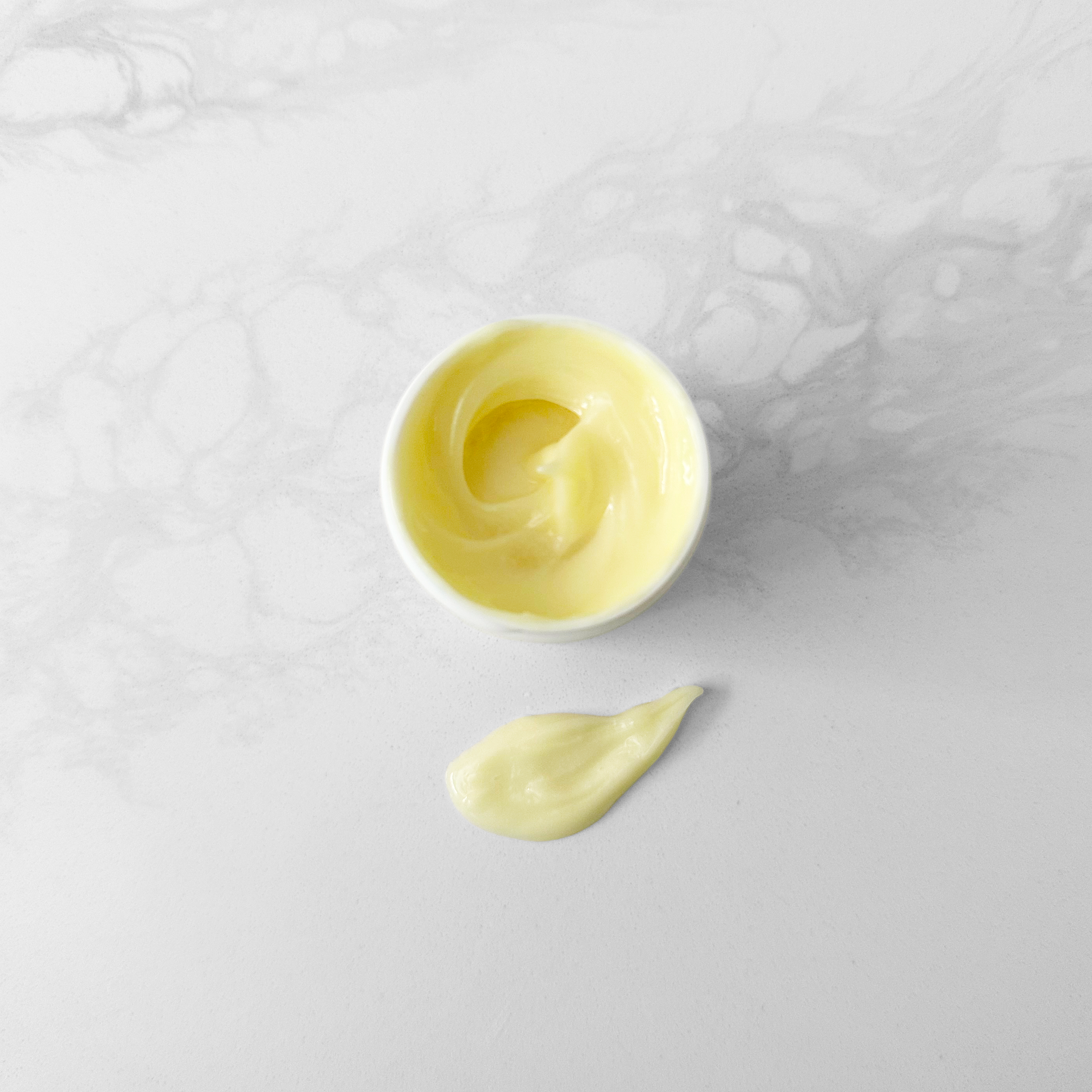 A small white jar containing pale yellow cream, with a dollop of the same product placed below it on a light gray marble-like surface.