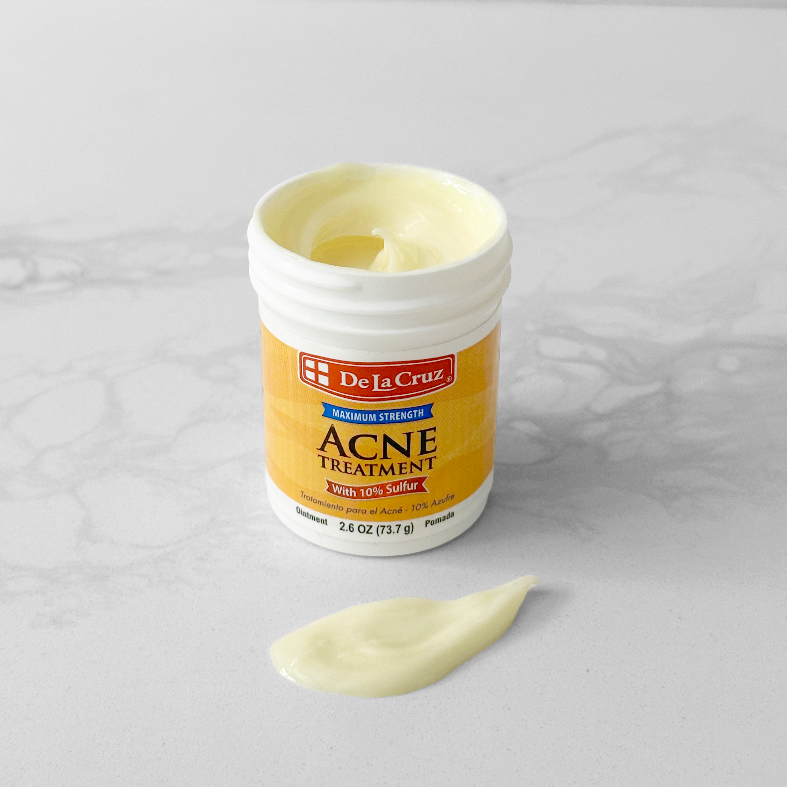 A white plastic jar of De La Cruz Acne Treatment with a yellow label, shown open with a dollop of pale yellow cream on a light gray marble surface.