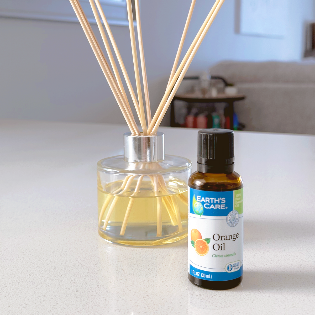 A reed diffuser bottle with clear glass, pale yellow liquid, and natural reed sticks sits on a white countertop next to a small amber bottle of Earth's Care Orange Oil, with a softly blurred indoor background.