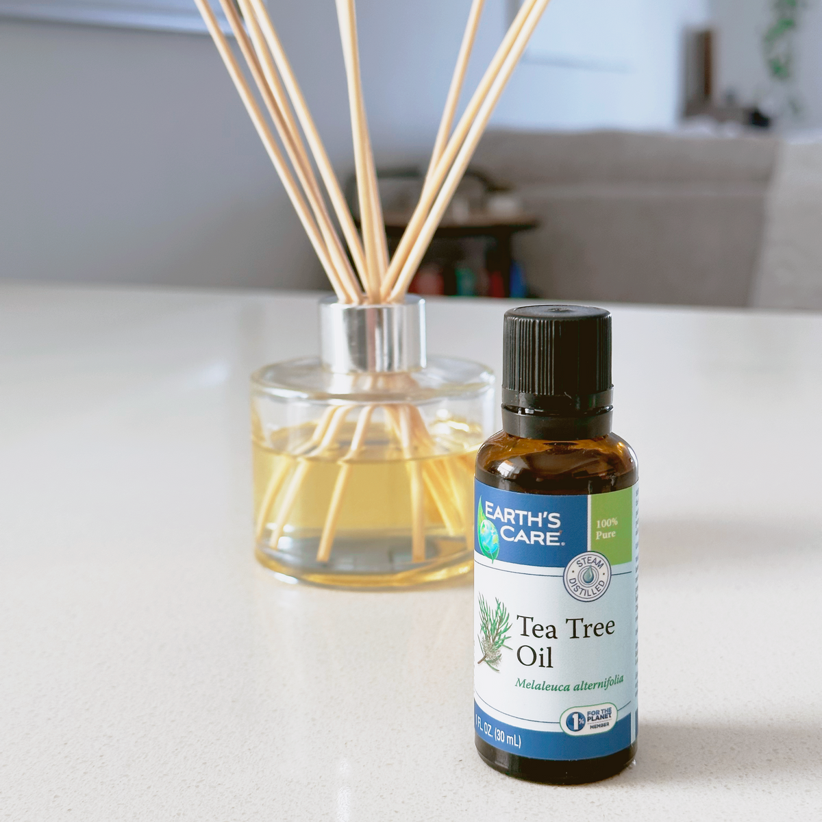 Earth's Care Tea Tree Oil bottle next to a reed diffuser with natural wood reeds on a light countertop in a blurred indoor setting.