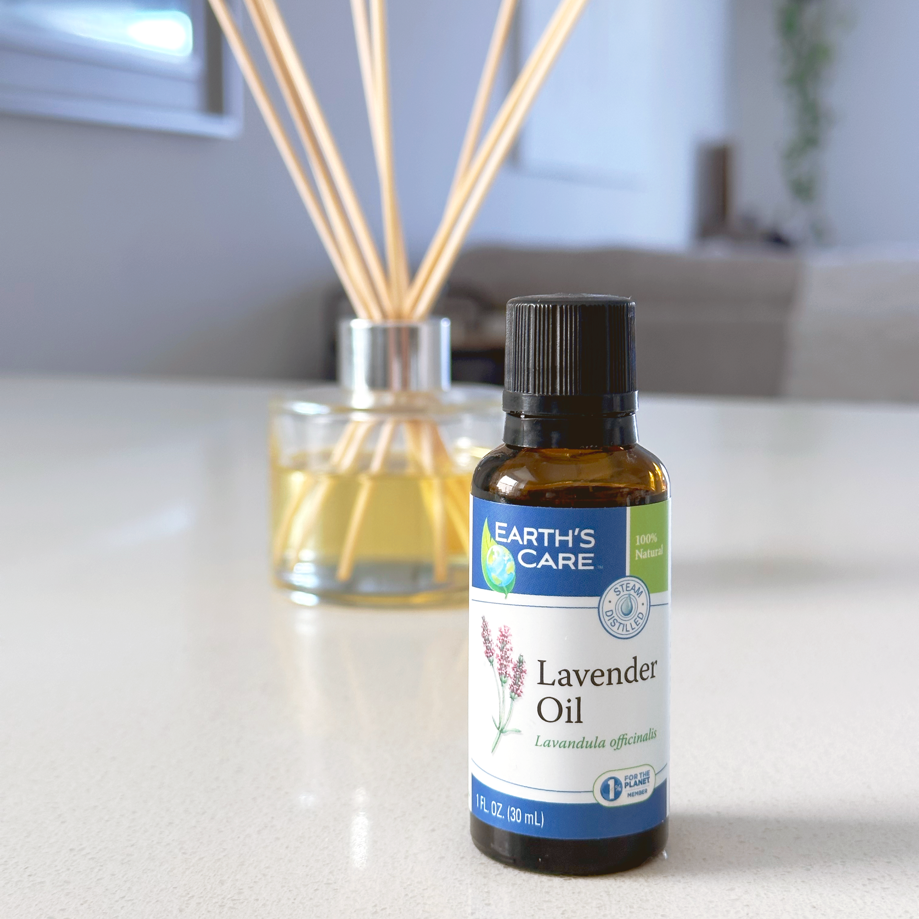A bottle of Earth's Care Lavender Oil in an amber glass bottle with a black cap, placed on a light countertop in front of a reed diffuser with wooden reeds in a softly blurred indoor setting.