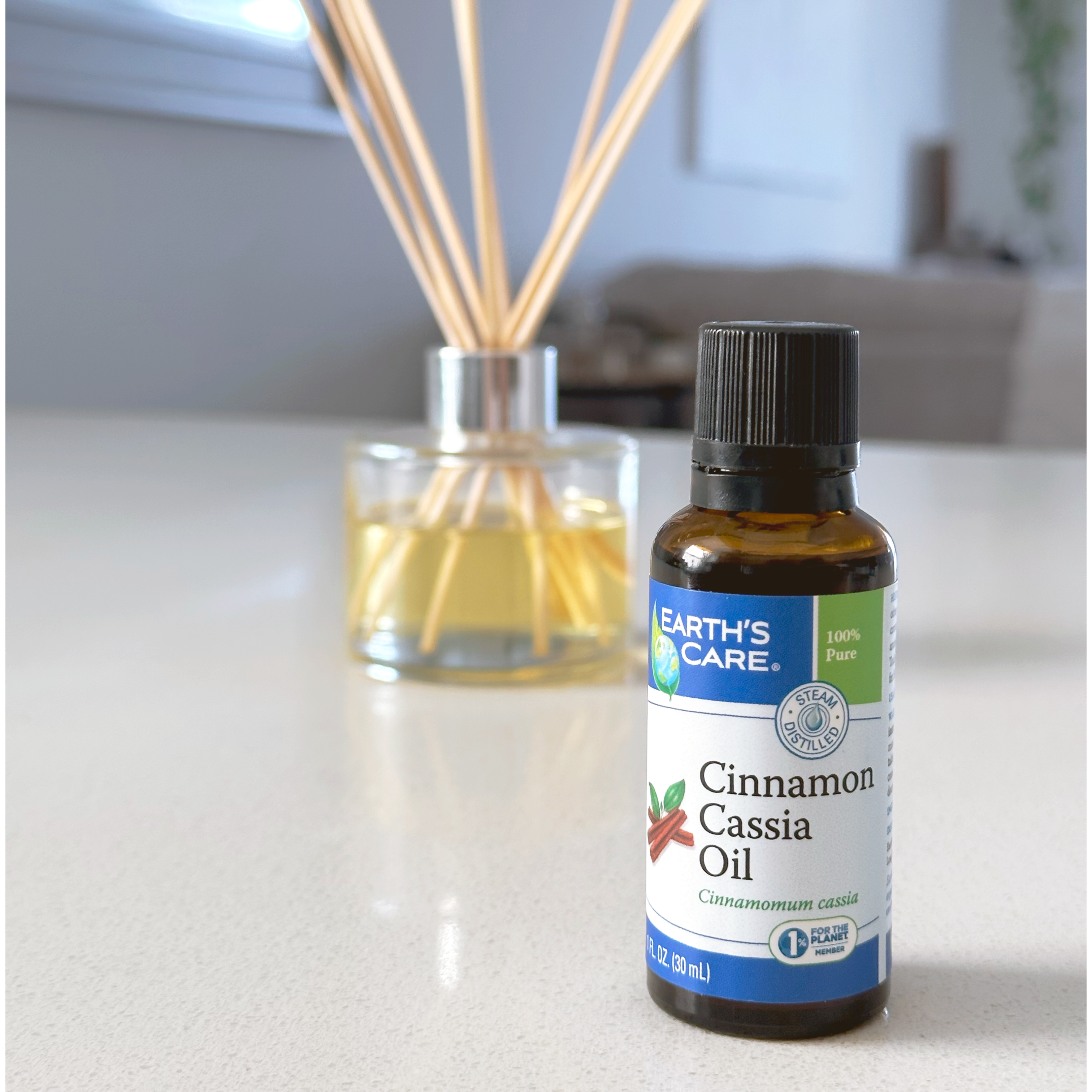A bottle of Earth's Care Cinnamon Cassia Oil in an amber glass bottle with a black cap, standing on a light countertop in front of a reed diffuser with wooden reeds in a softly blurred indoor setting.