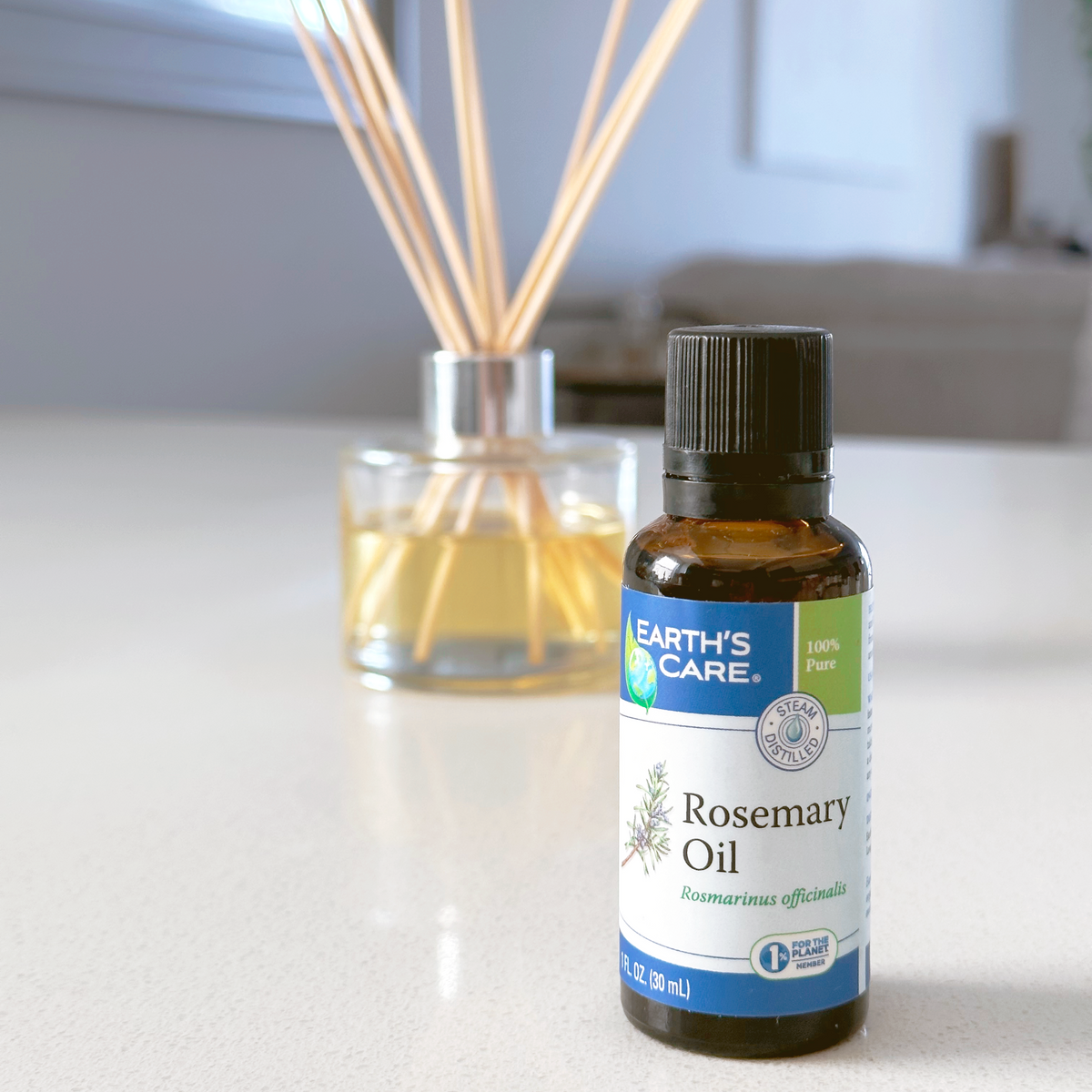 A bottle of Earth's Care Rosemary Oil essential oil in the foreground, with a glass reed diffuser in the blurred background on a light countertop.