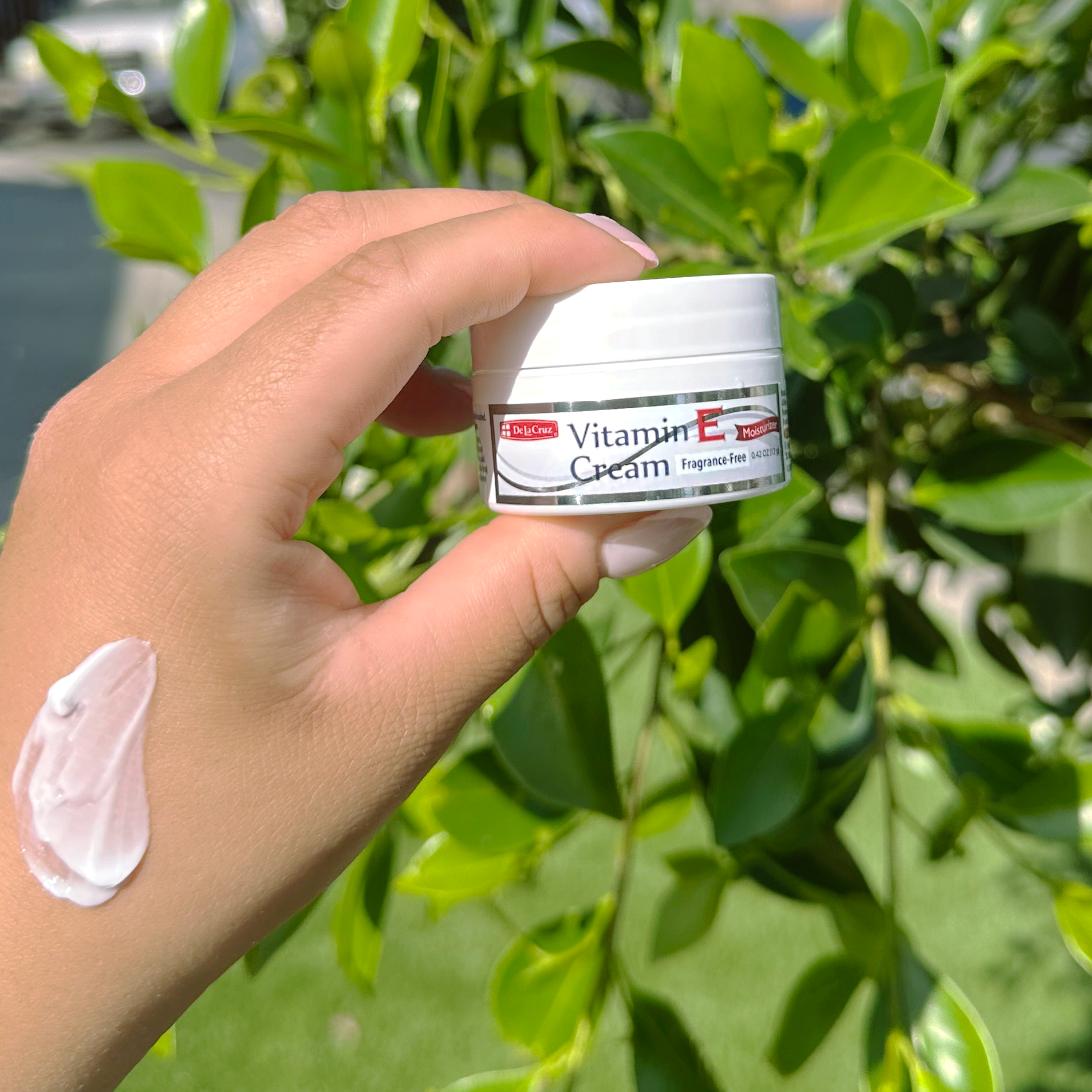 A hand holding a small white jar labeled Vitamin E Garden outdoors, with a smear of white cream on the wrist and green foliage in the background.