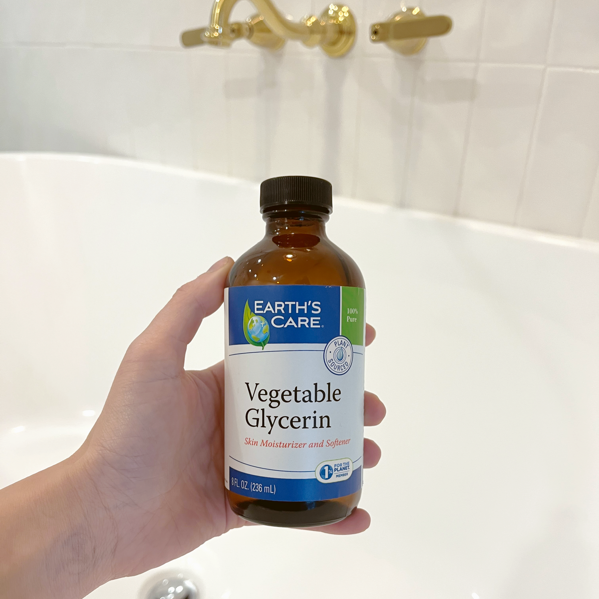 A hand holds an amber glass bottle of Earth's Care Vegetable Glycerin in front of a white bathtub with gold wall-mounted faucets.