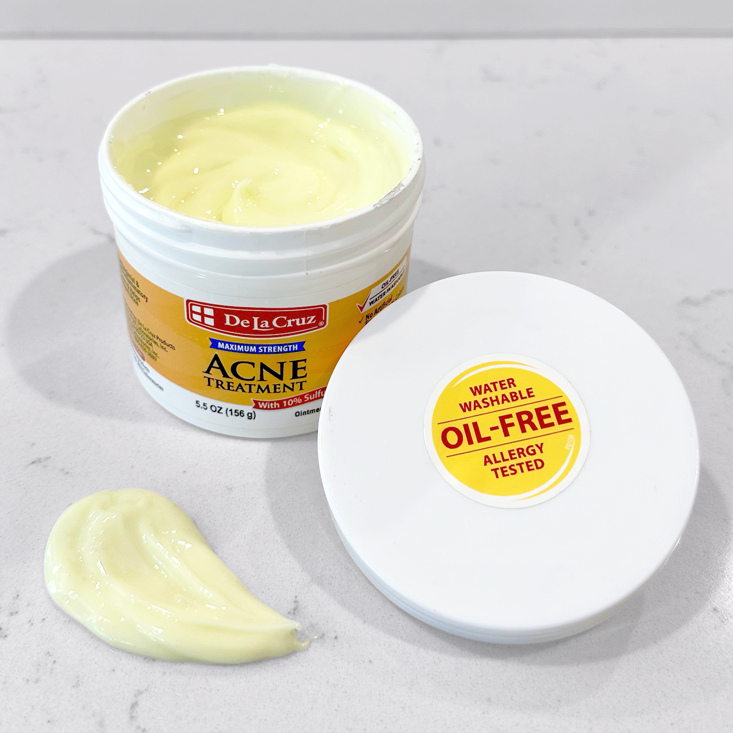 A white plastic jar of De La Cruz Acne Treatment with pale yellow cream visible inside, shown next to its lid and a dollop of the ointment on a light marble surface.